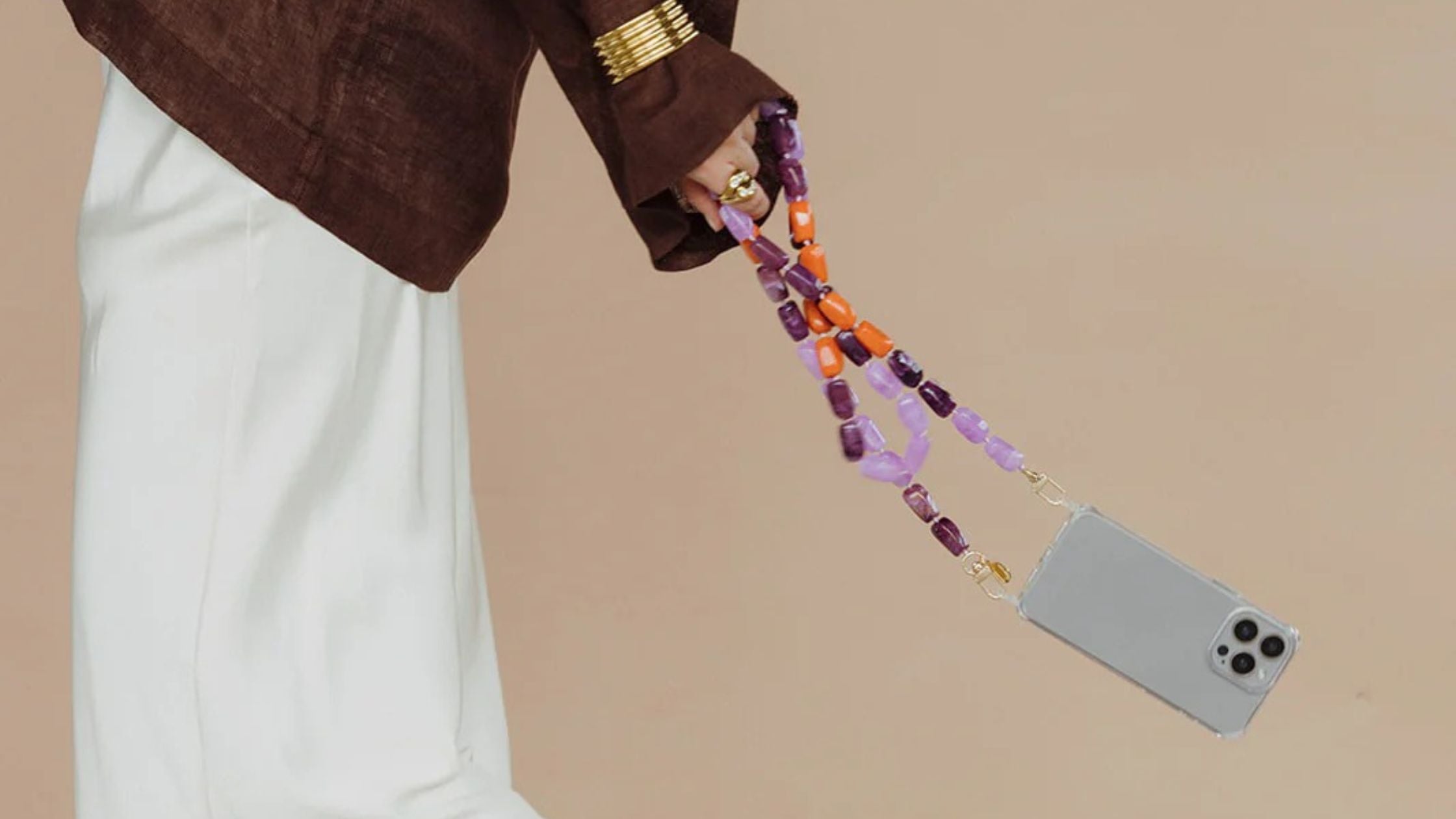 Woman's hand holding a phone with a colorful beaded phone charm strap attached, wearing a brown jacket.