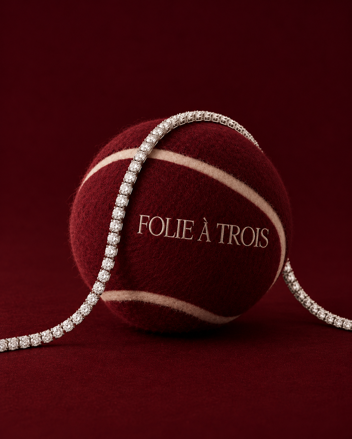 Silver-tone Rivière tennis NECKLACE draped over a burgundy tennis ball printed with Folie à Trois.