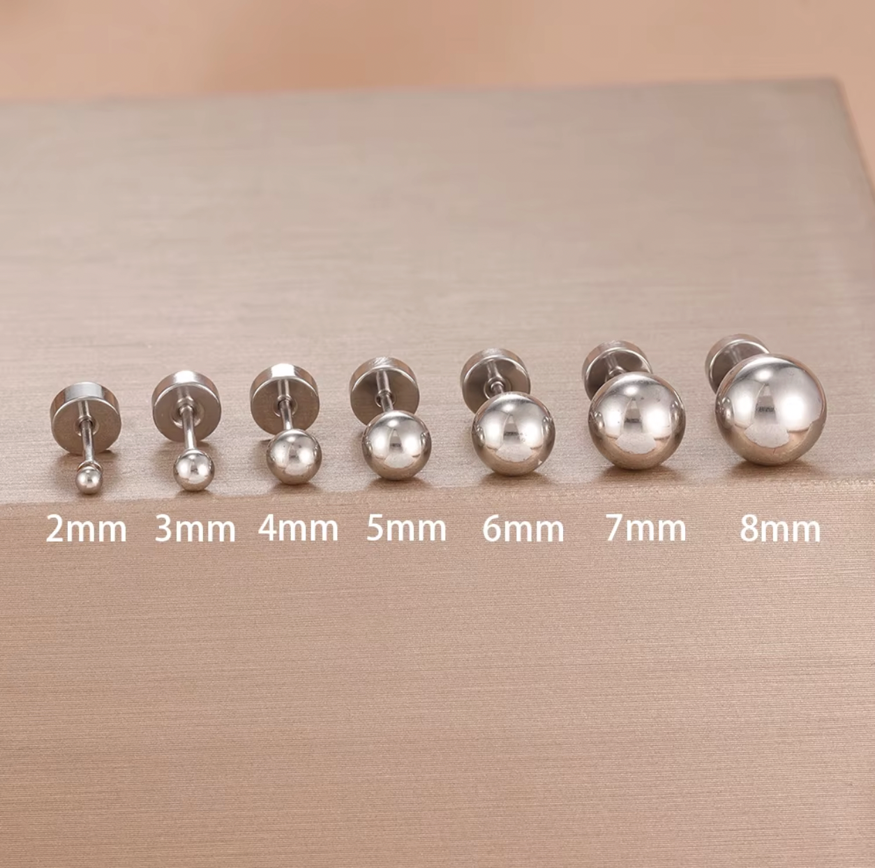 Row of gold ball stud piercings in multiple sizes displayed on a soft fabric background, showing smooth polished finish and flat screw-back closures.