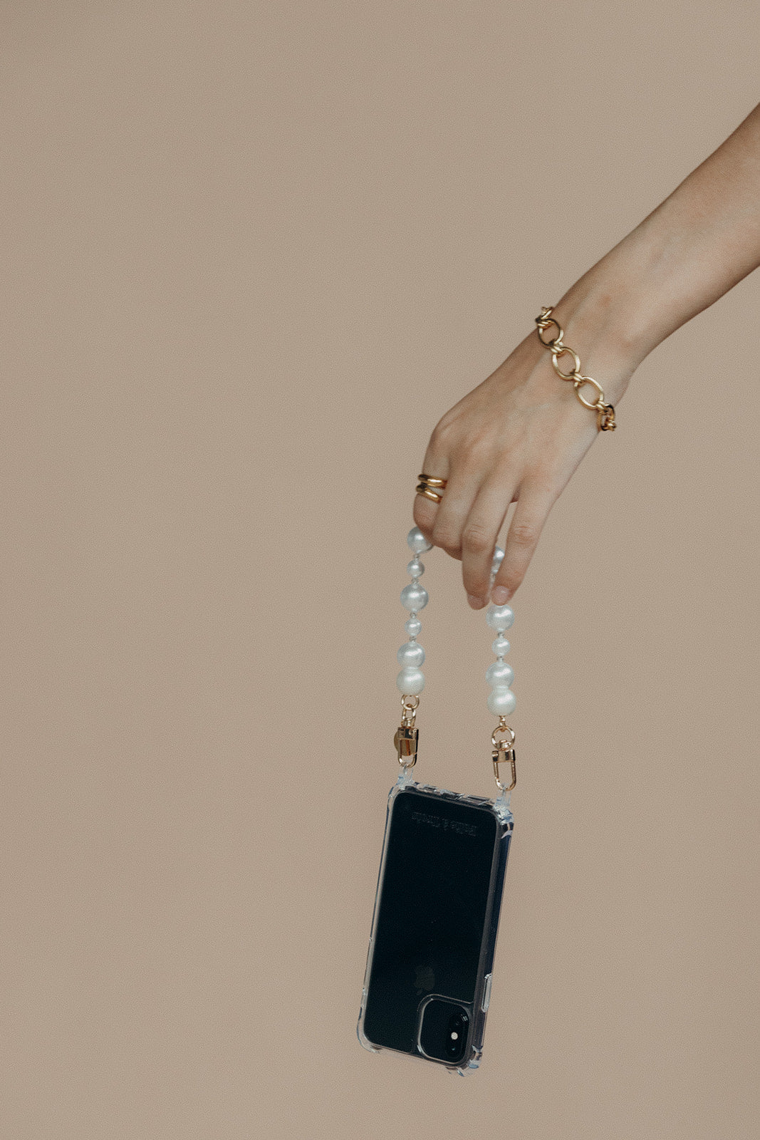 Model's hand and gold bracelet holding a phone in a clear case suspended from the Folie à Trois Adair Pearl wholesale phone chain.