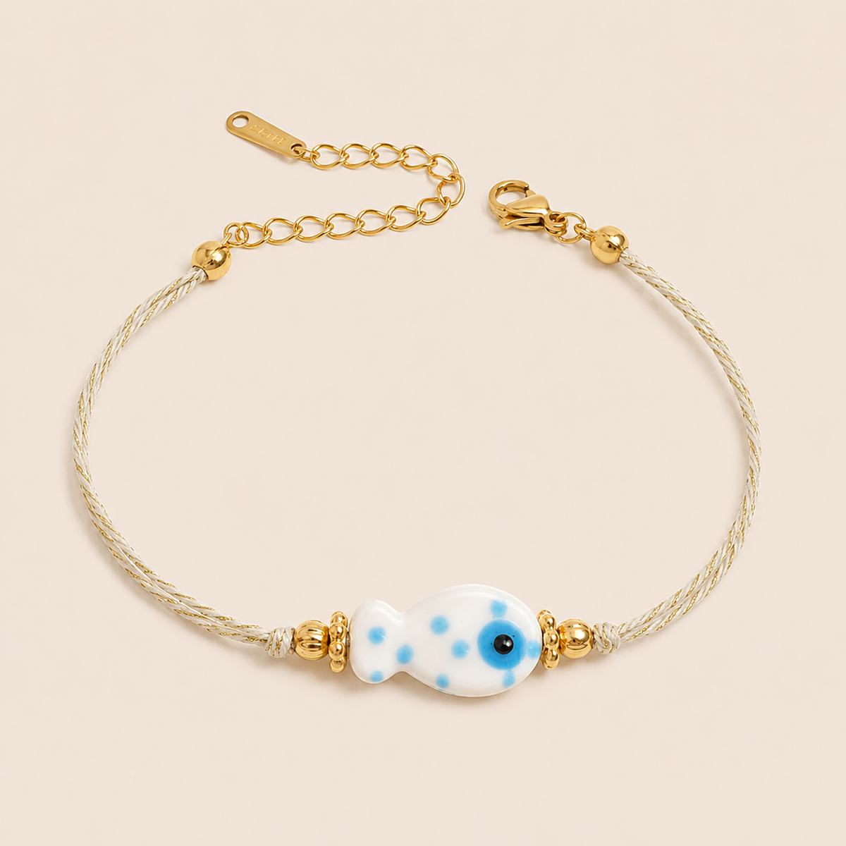Amalfi BRACELET in white with a ceramic blue polka-dot fish charm on a twisted gold and cream cord, shown flat.