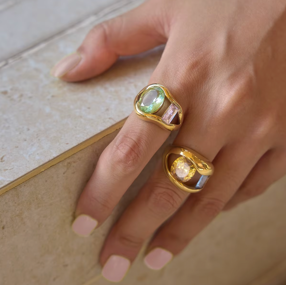 Model rests her hand on a stone ledge wearing two Amara RING colorways stacked, green-pink and yellow-blue gemstone pairings visible.