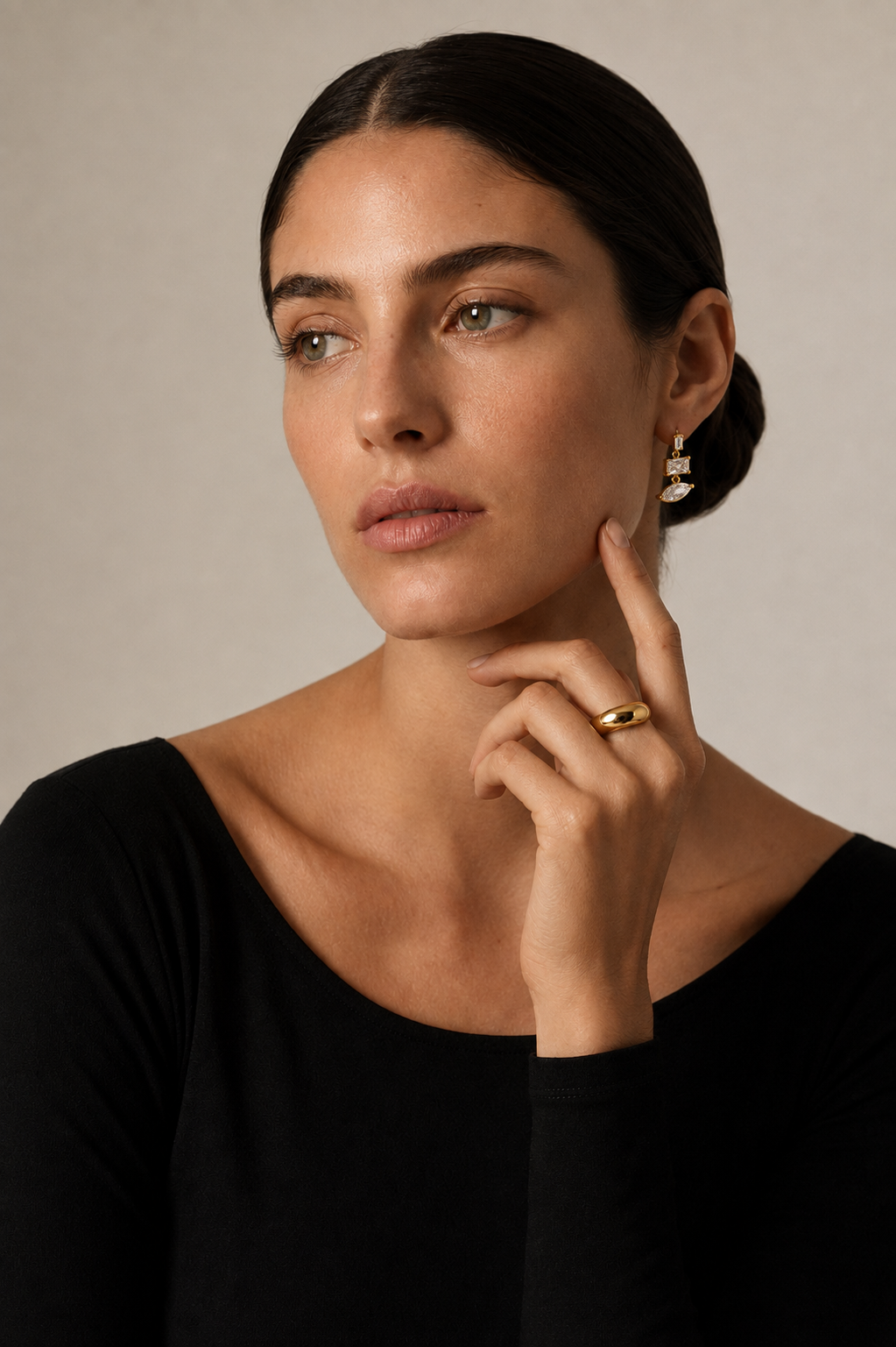 Model in black touches her cheek, wearing the gold Angles EARRINGS kite and marquise zirconia drop with a chunky gold ring.