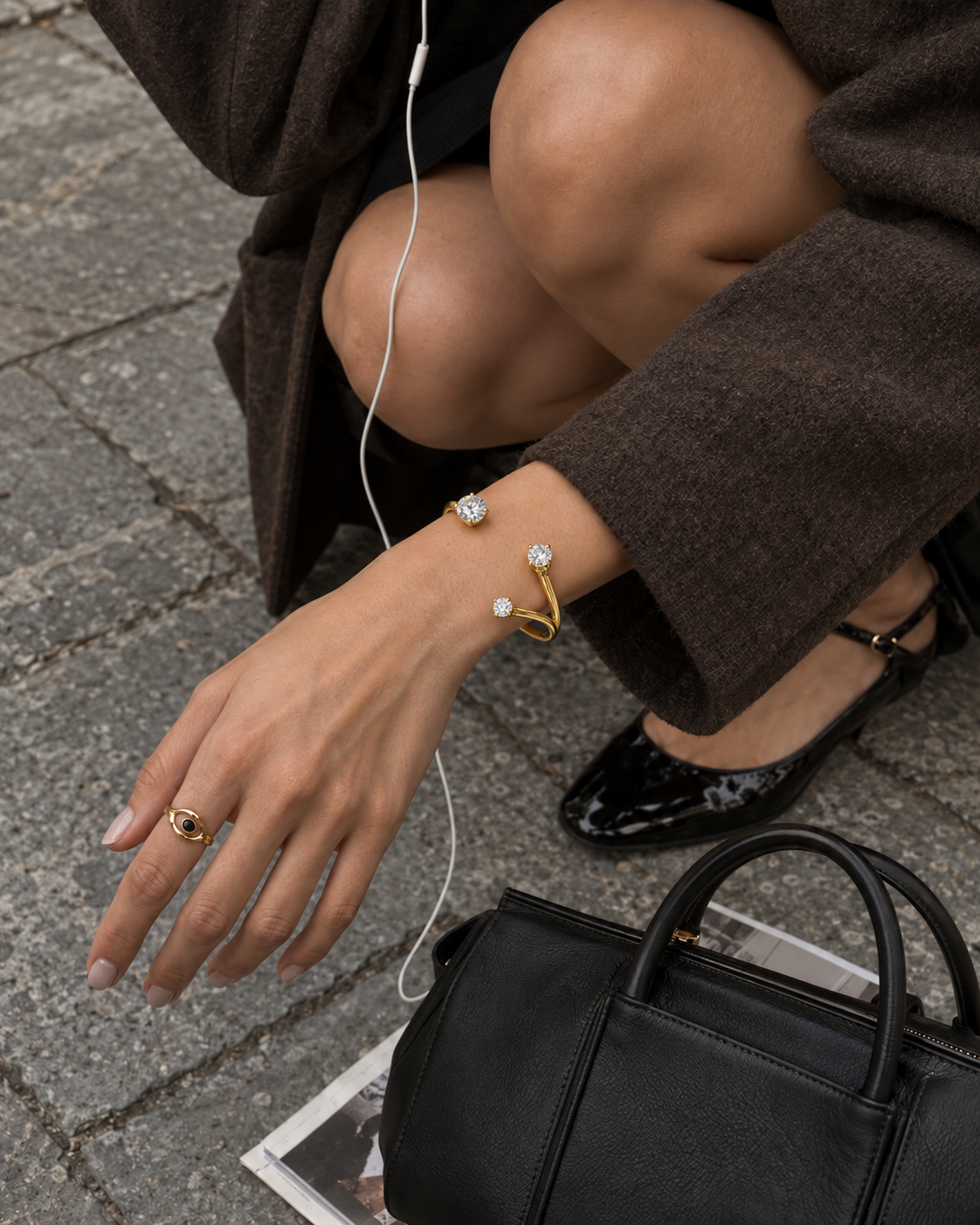 Crouching model in a brown coat wears the gold Aurelia BRACELET cuff with zirconia ends beside a black handbag — Folie à Trois