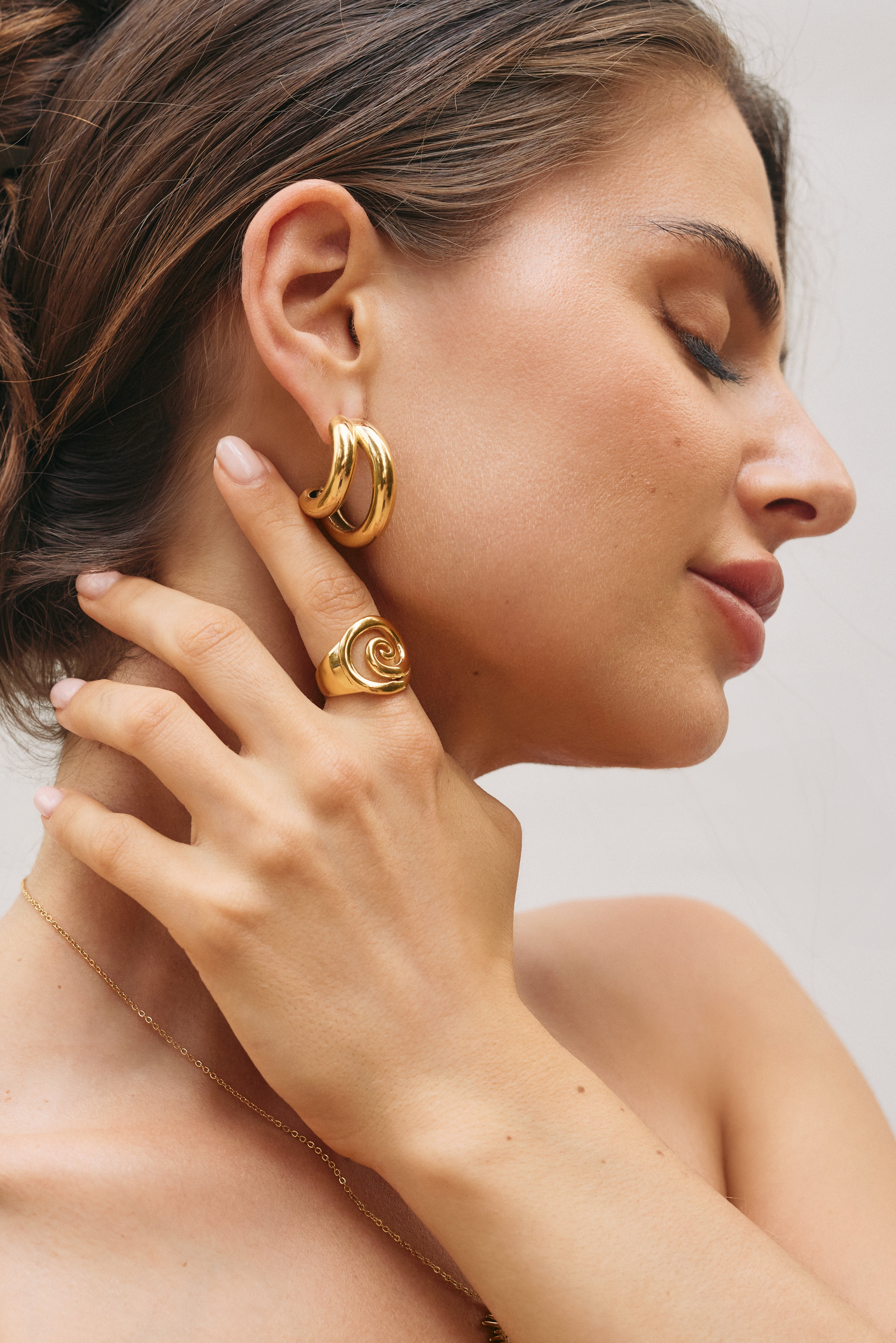 Gold signet-style ring with a spiral swirl carved into the wide oval face, shown at an angle on a cream backdrop — Avery RING.