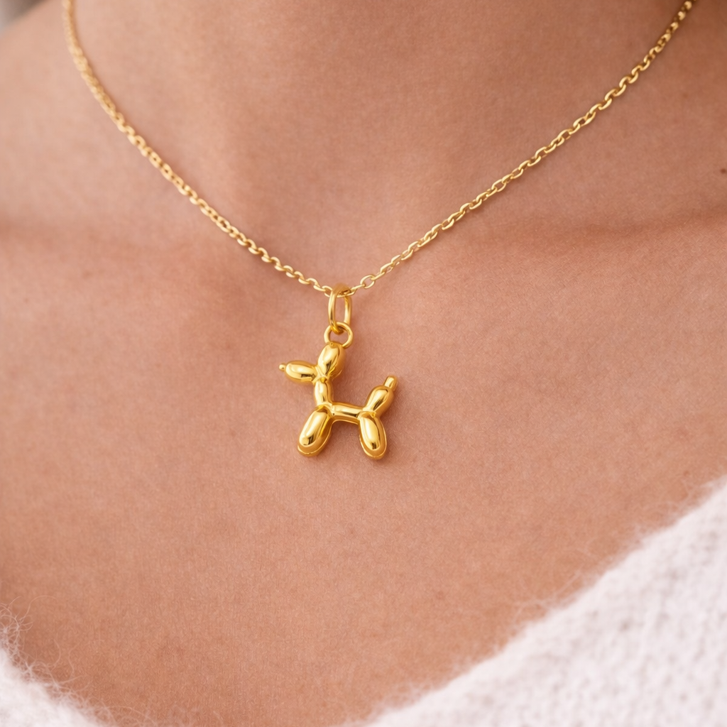 The Balloon Dog CHARM worn at the neckline, a polished gold balloon-animal dog pendant on a fine gold chain against a cream sweater.