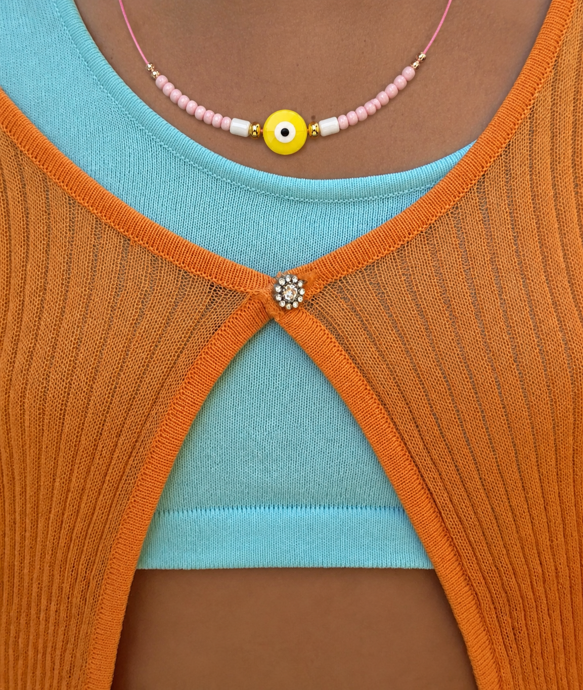 Model wears the Big Eye NECKLACE close-up, a yellow enamel evil eye on a pink beaded cord at the neckline.