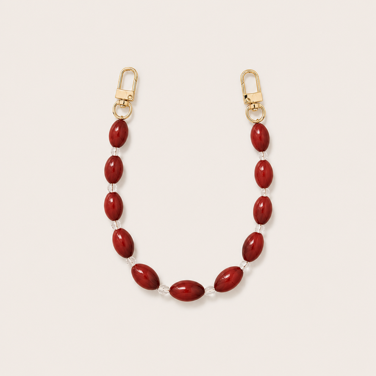 Folie à Trois Bramble short wholesale phone chain in deep red oval beads with clear glass spacers and gold clasps, laid flat on a cream background.