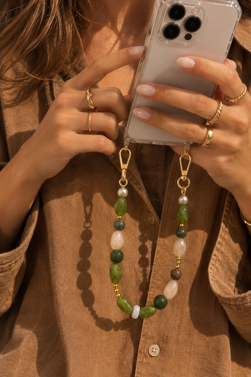 Model in a tan shirt holding a clear phone case attached to the Folie à Trois Briar wholesale phone chain in mixed green and blush beads.