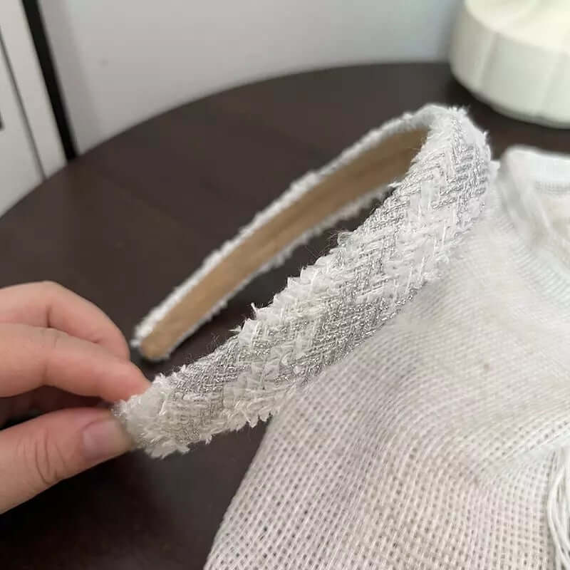 A single cream and silver metallic diamond-weave tweed headband held by hand on a dark table.
