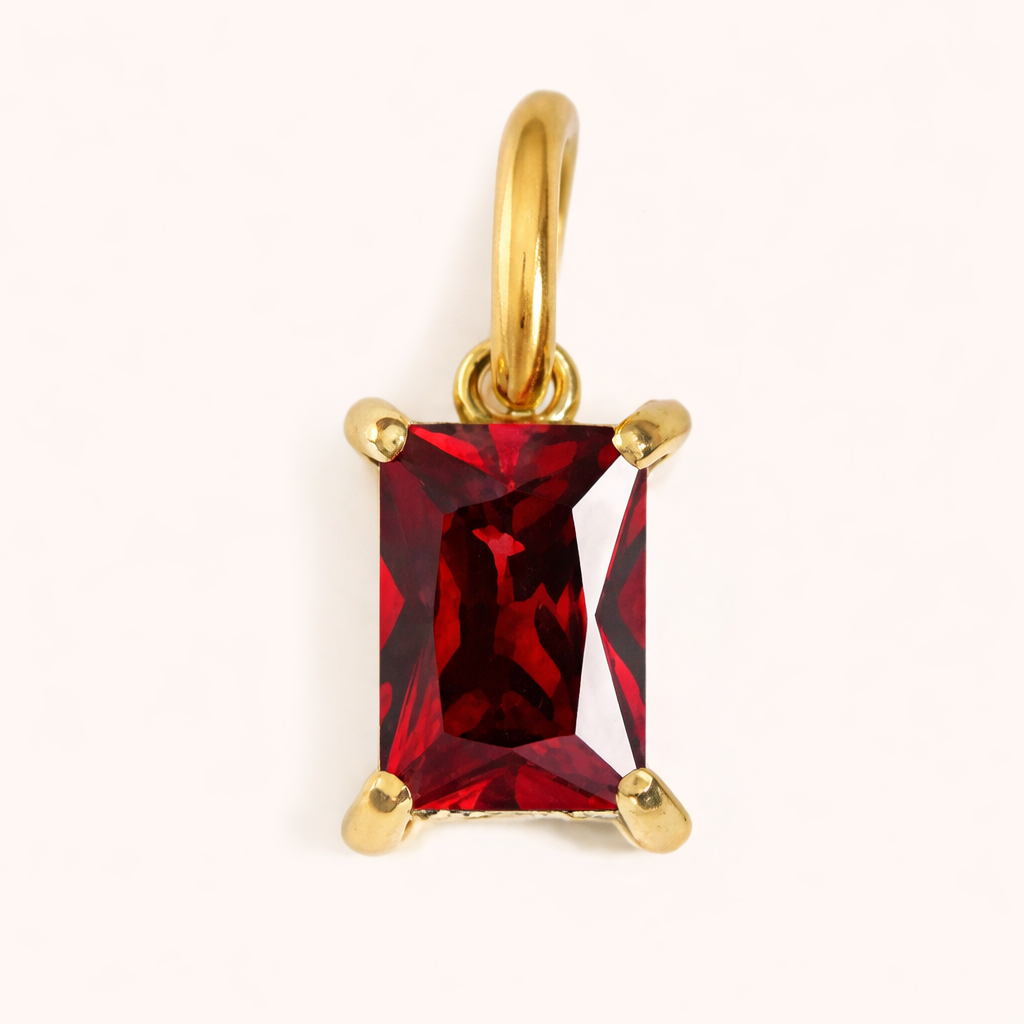Close-up of the Classic Drop Charm in a rich red garnet stone, set in a gold prong mount on a cream background.