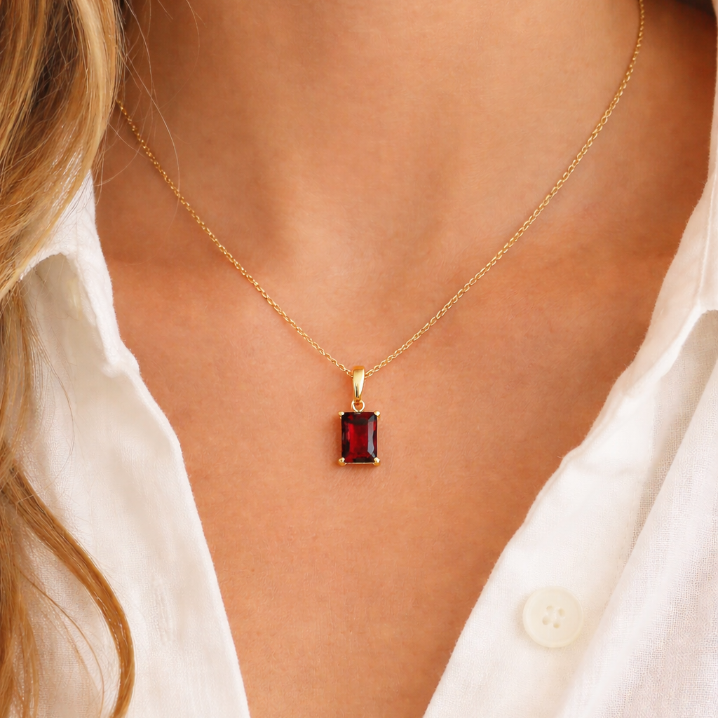 A Classic Drop Charm in deep red garnet hangs from a slim gold chain necklace, worn against skin beneath an open white blouse collar.