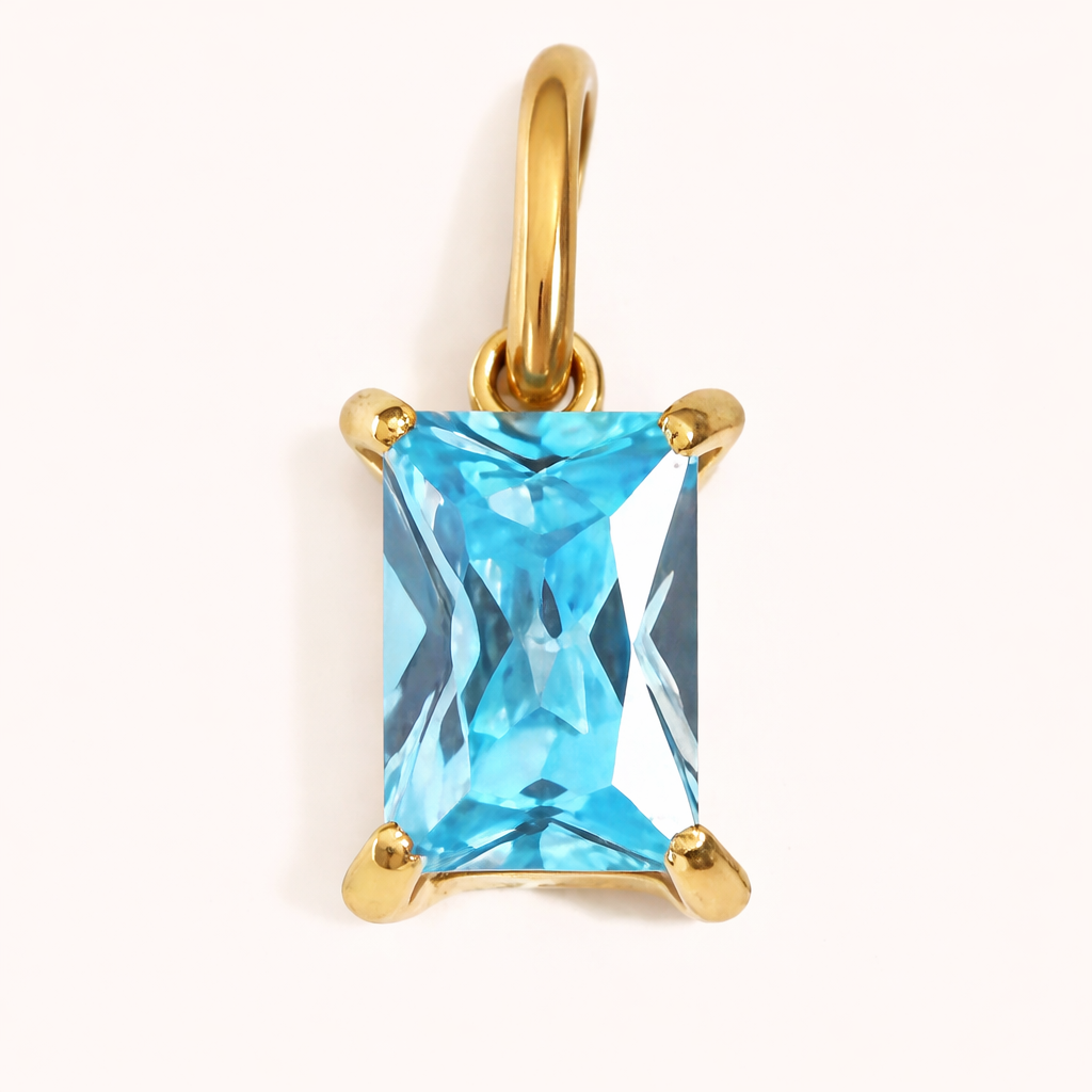 Close-up of the Classic Drop Charm in a bright aqua blue stone, set in a gold prong mount on a cream background.