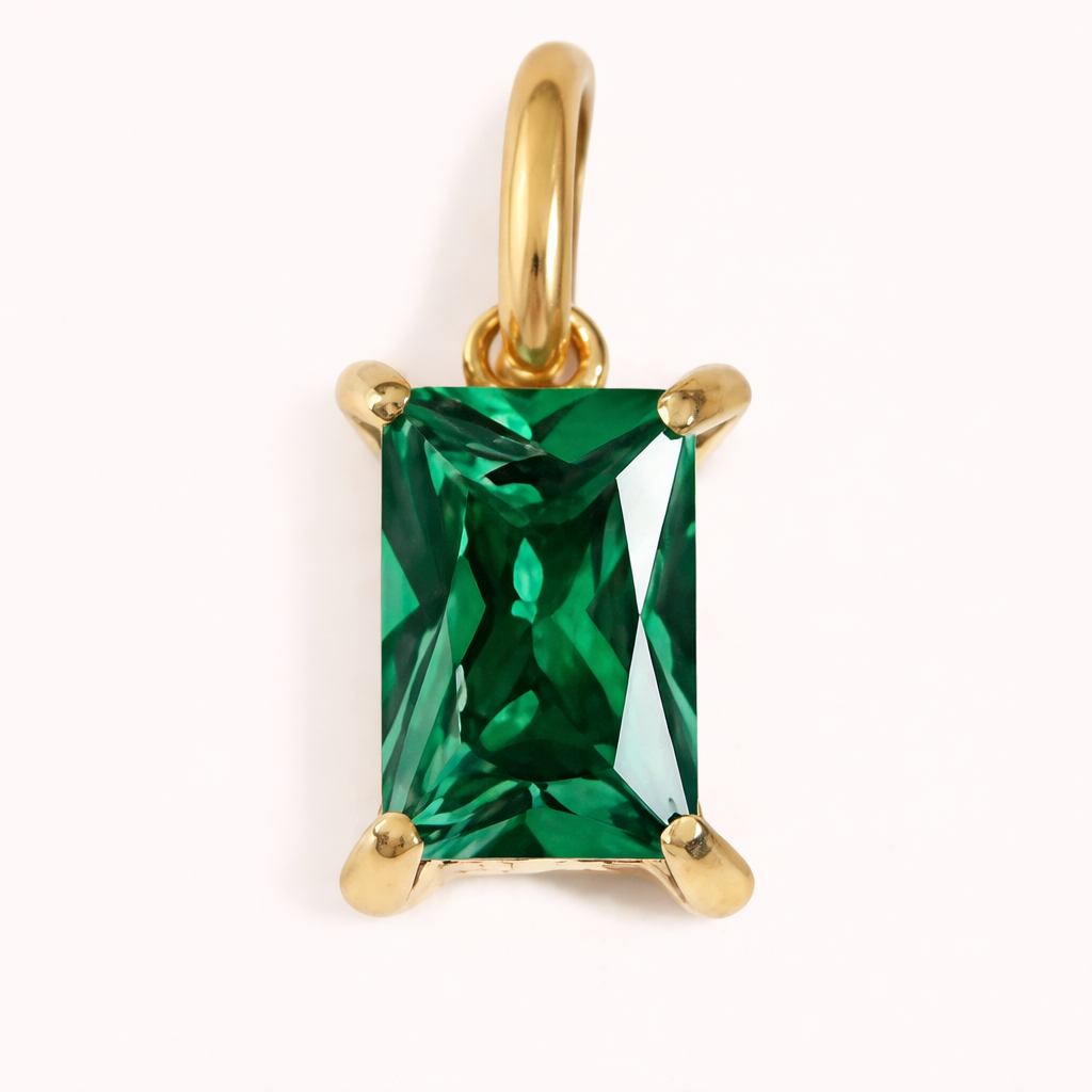 Close-up of the Classic Drop Charm in a vivid emerald green stone, set in a gold prong mount on a cream background.