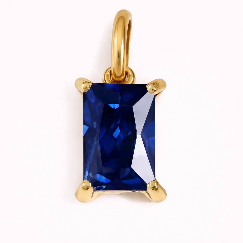 Close-up of the Classic Drop Charm in a deep sapphire blue stone, set in a gold prong mount on a cream background.