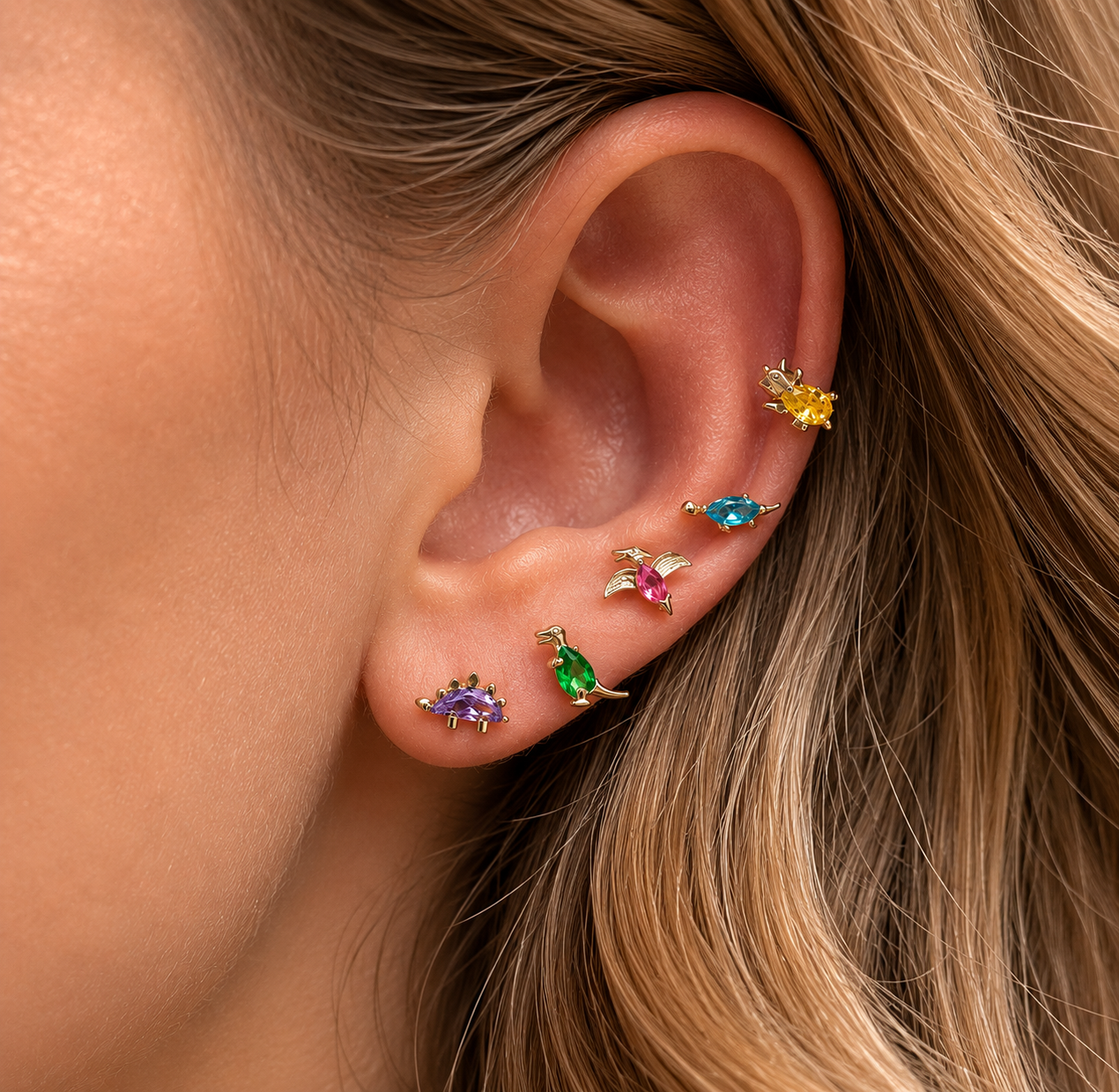 Close-up of a model's ear stacked with five tiny gold dinosaur studs from the Colorful Dino EARRINGS in purple, green, pink, and yellow.