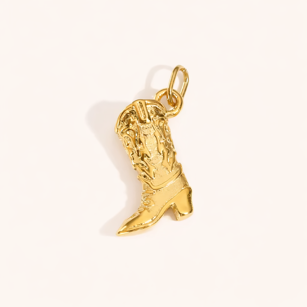 The Cowboy CHARM in a studio flat lay, a textured gold pendant shaped like a western cowboy boot on a cream background.