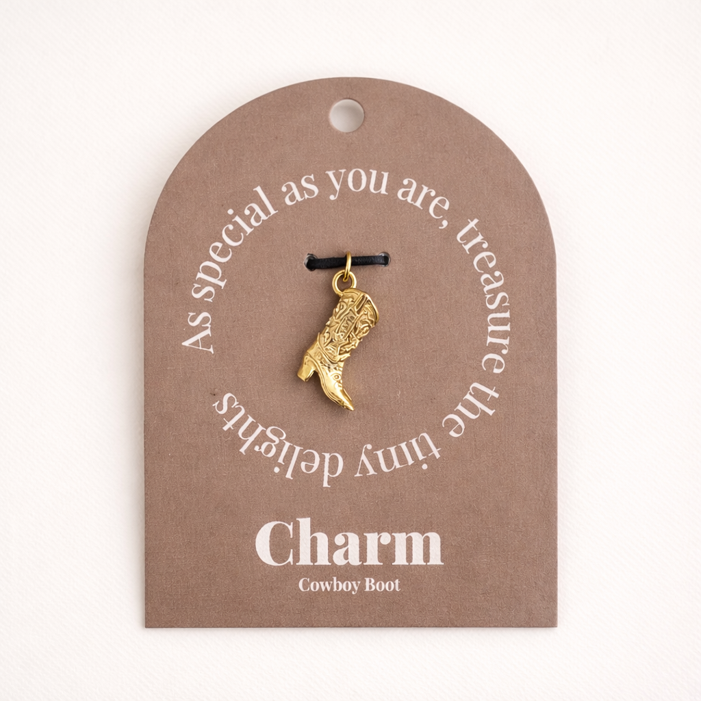 The Cowboy CHARM displayed on a tan Charm gift tag, the textured gold cowboy boot pendant hanging from a black cord.