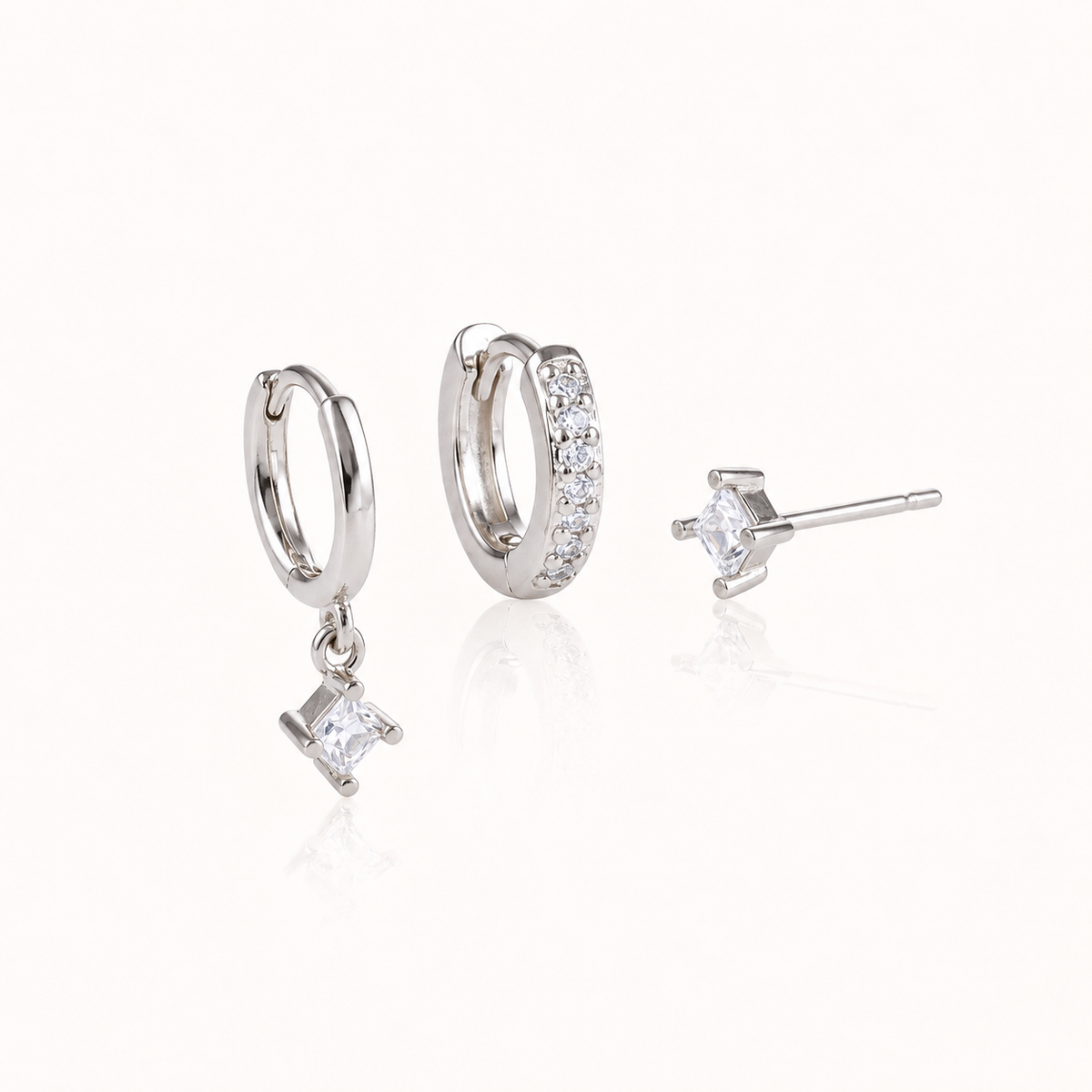 Three silver Eira SET pieces: a huggie with a square crystal drop, a pavé huggie hoop and a square crystal stud, on white.