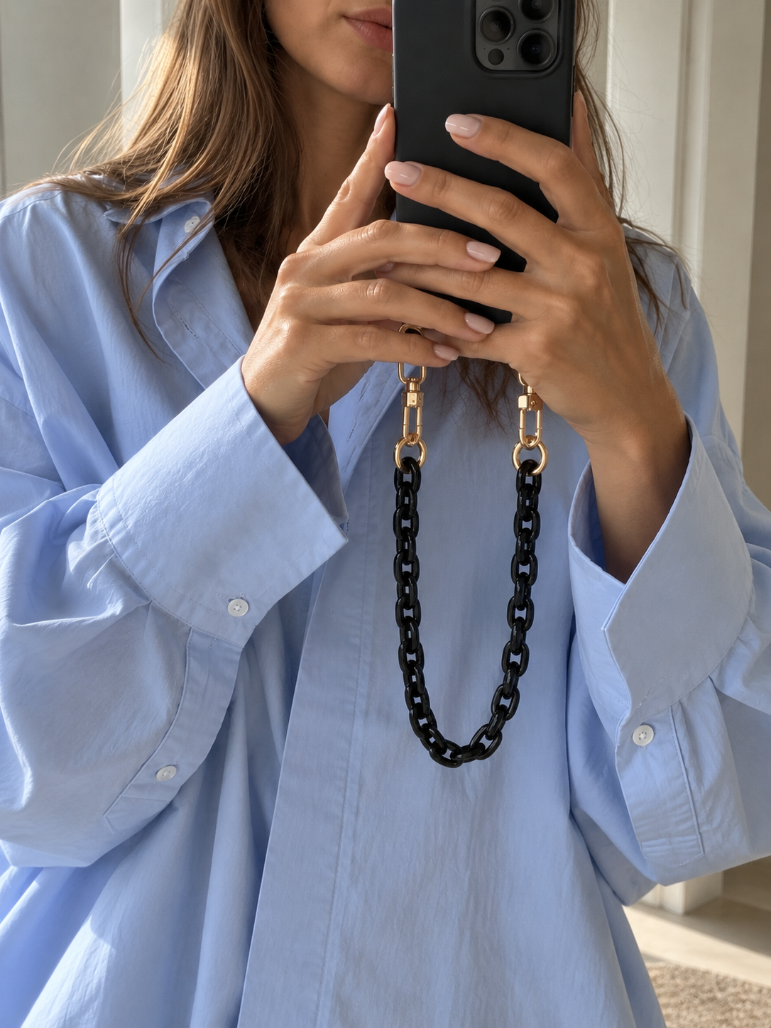 The Folie à Trois Ember SHORT PHONE CHAIN in glossy black acrylic, clipped to a phone held up for a mirror selfie in a light blue shirt.