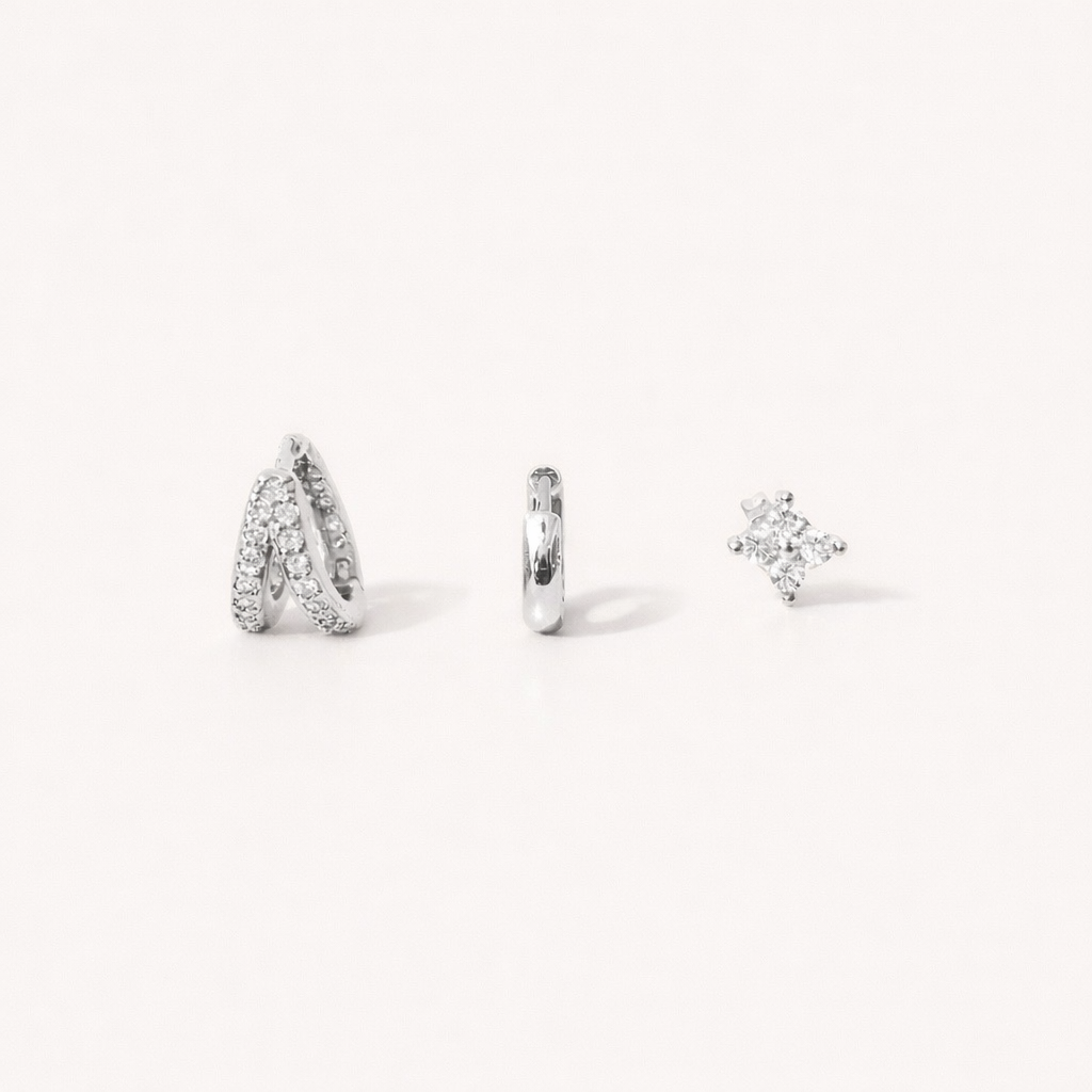 Faye Set in silver tone, a pave crossover hoop, a plain huggie hoop, and a four-point crystal stud, laid flat.