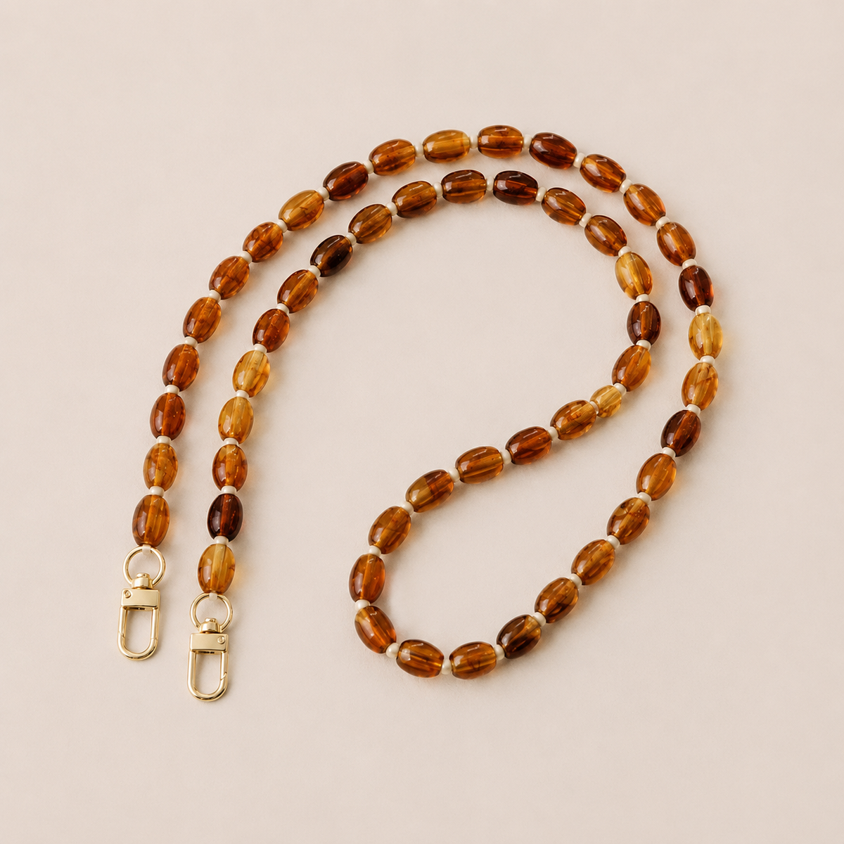 Folie à Trois Fennel long phone chain in amber oval beads with cream spacers and gold clasps, laid flat on a cream background.