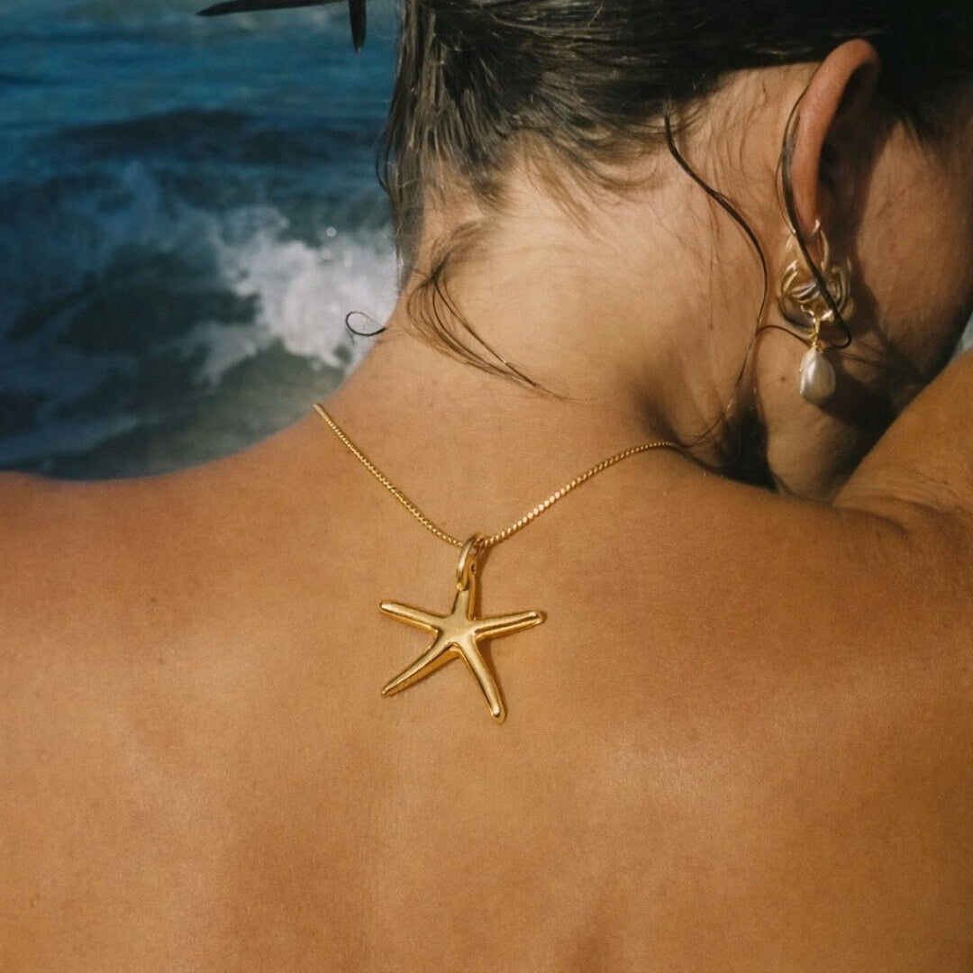 The Fiji NECKLACE worn from behind at the nape of the neck, its gold starfish resting against tanned skin with the ocean behind.