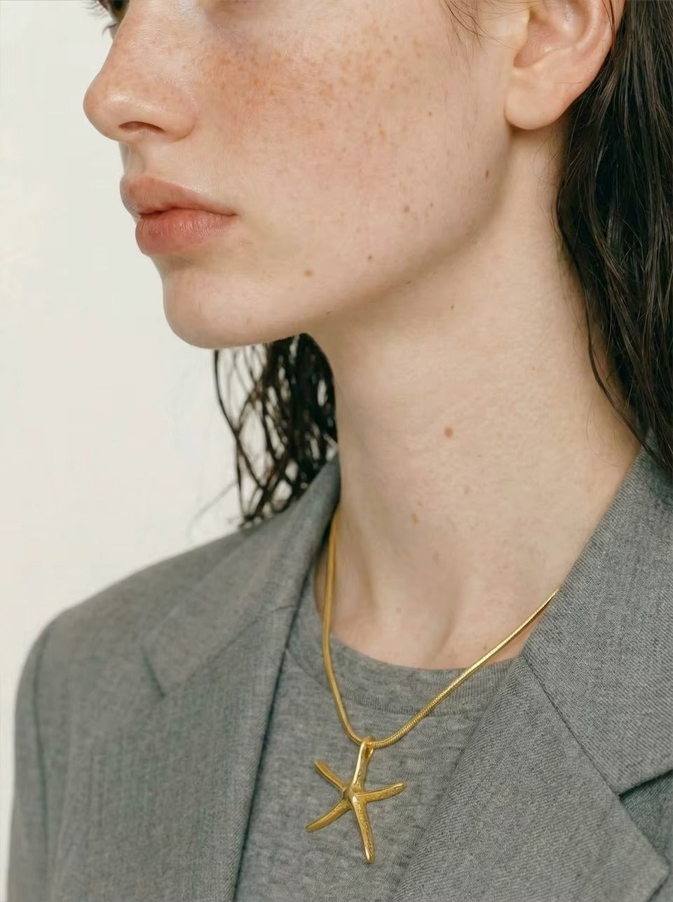 The Fiji NECKLACE worn in profile on a gray tee, the gold starfish resting at the collarbone beneath a layered chain.