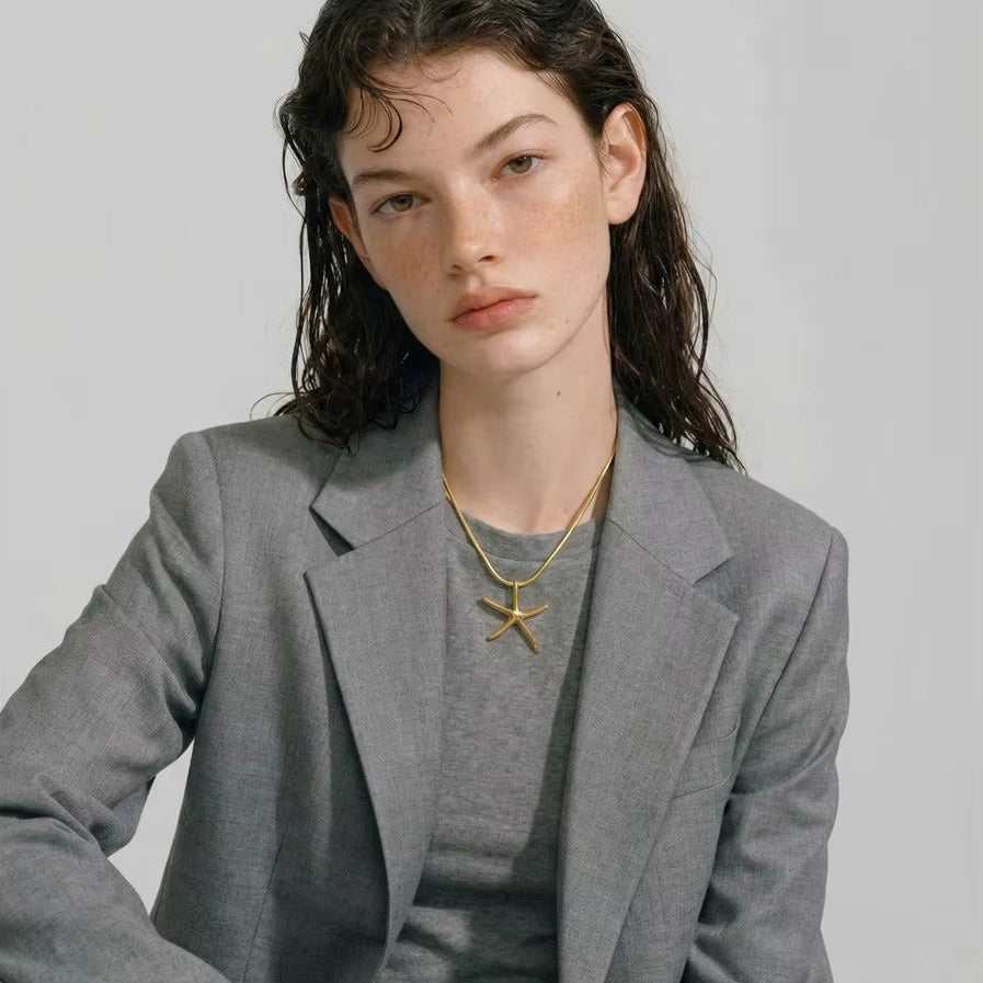 The Fiji NECKLACE worn over a gray blazer and tee, the model seated and looking directly at the camera.