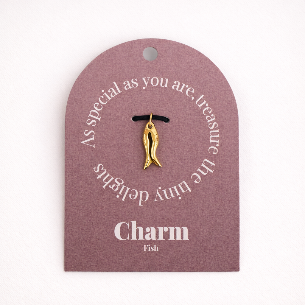 The Fish CHARM displayed on a mauve Charm gift tag, the sculptural gold fish pendant hanging from a black cord.