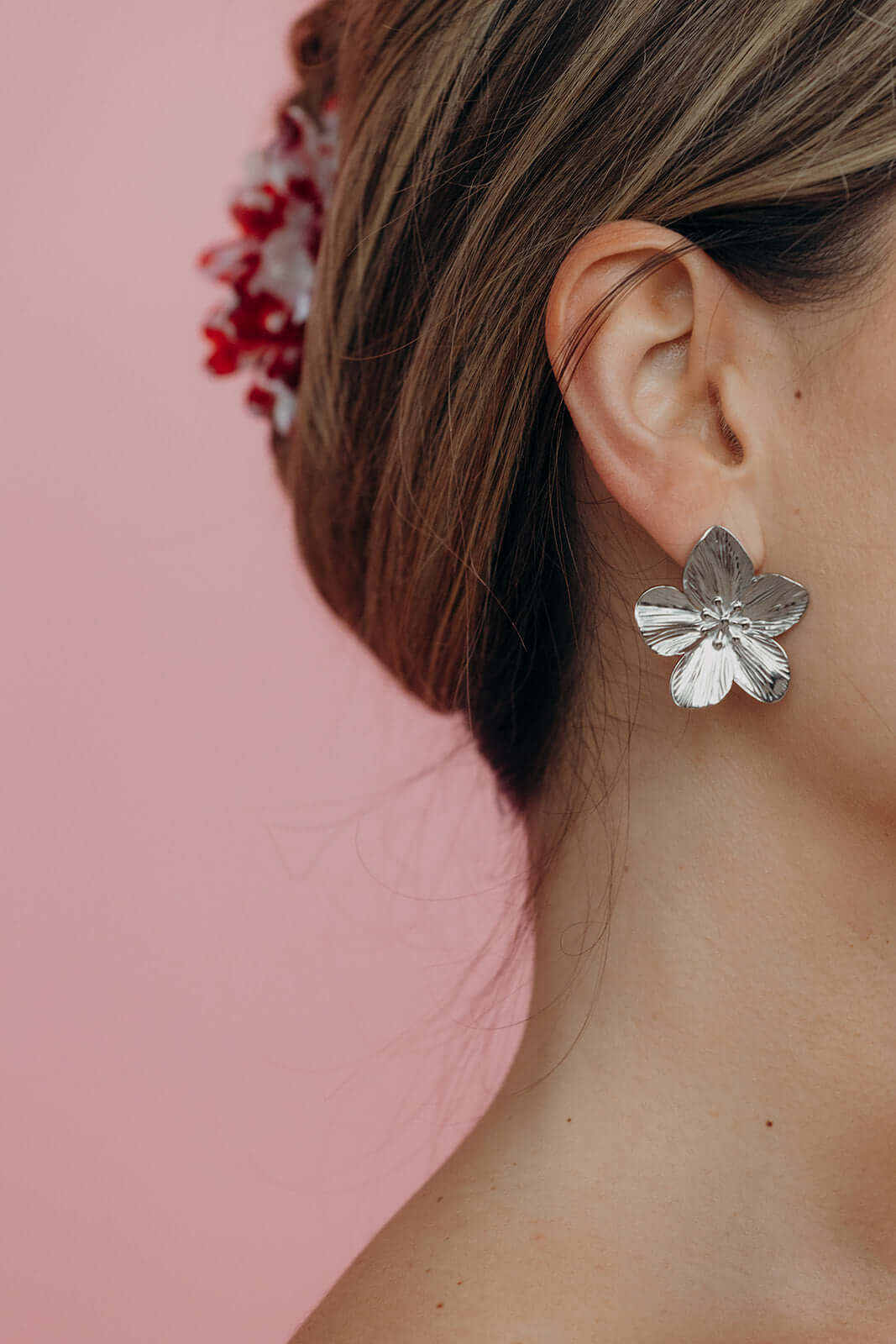 Woman with a low bun wearing the Funky HAIR CLIP with red flowers and a silver flower earring, against a pink wall.