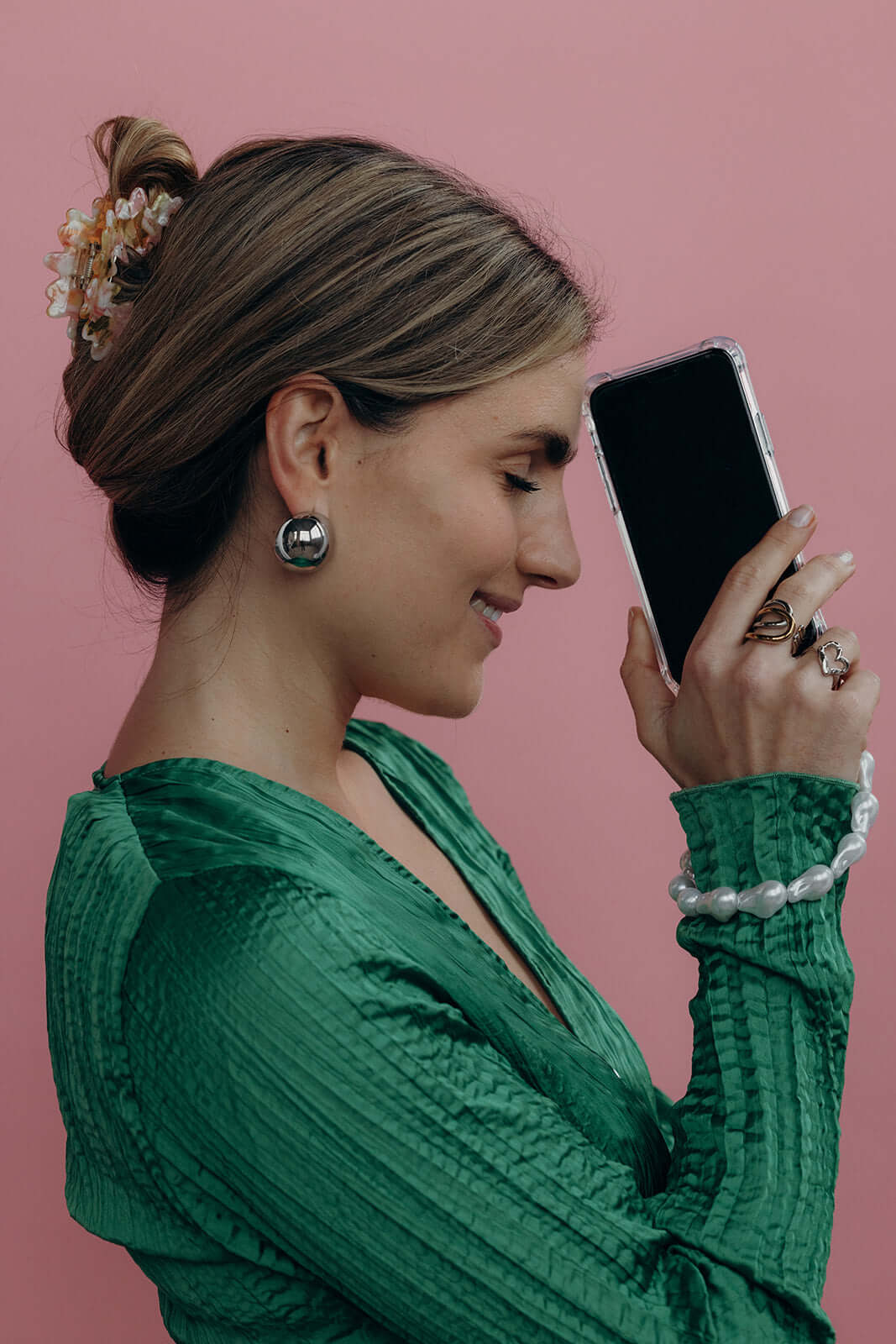 Profile of a woman in a green wrap blouse with a floral Funky HAIR CLIP in her bun, smiling at her phone on a pink wall.