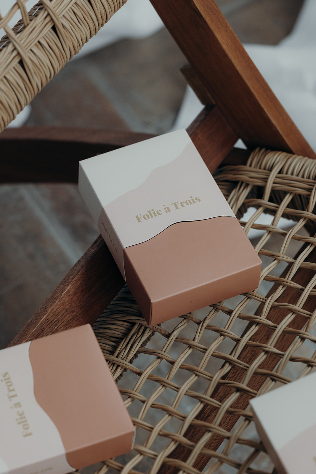 Folie à Trois Gift Box in cream and terracotta with a gold logo, resting on a woven rattan chair with more boxes nearby.