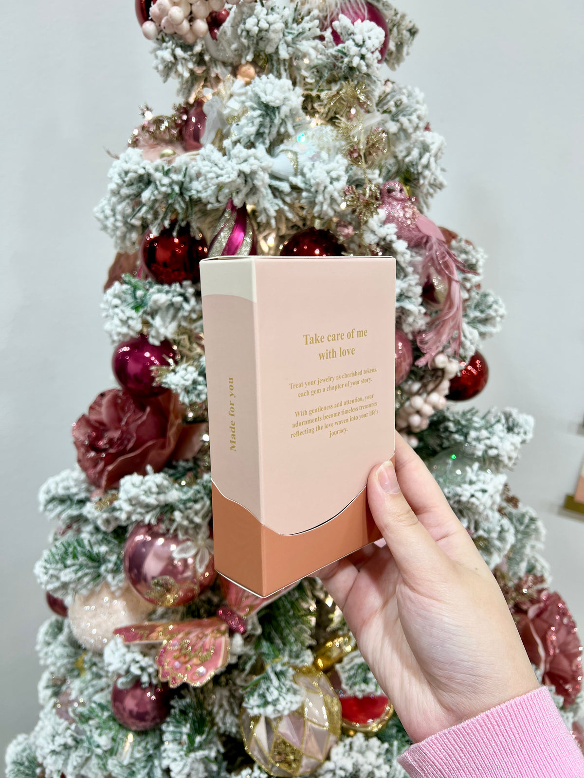 Hand holding the back of a cream and terracotta Folie à Trois Gift Box in front of a flocked Christmas tree with pink baubles.
