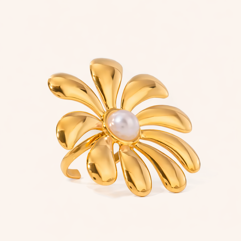 Gold ring shaped like a spiraling flower with curved petals and a pearl center, shown at an angle on a cream backdrop — Glistening Embrace RING.