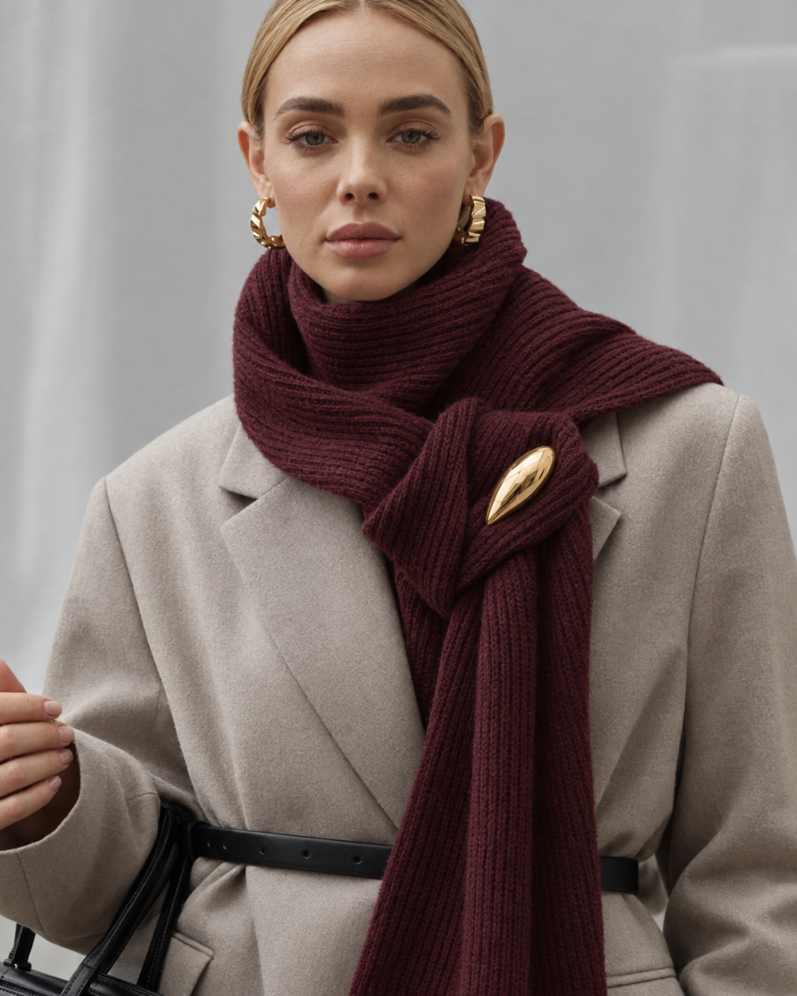 A woman wearing a chunky ribbed burgundy scarf with a large gold teardrop pin, styled over a beige coat.