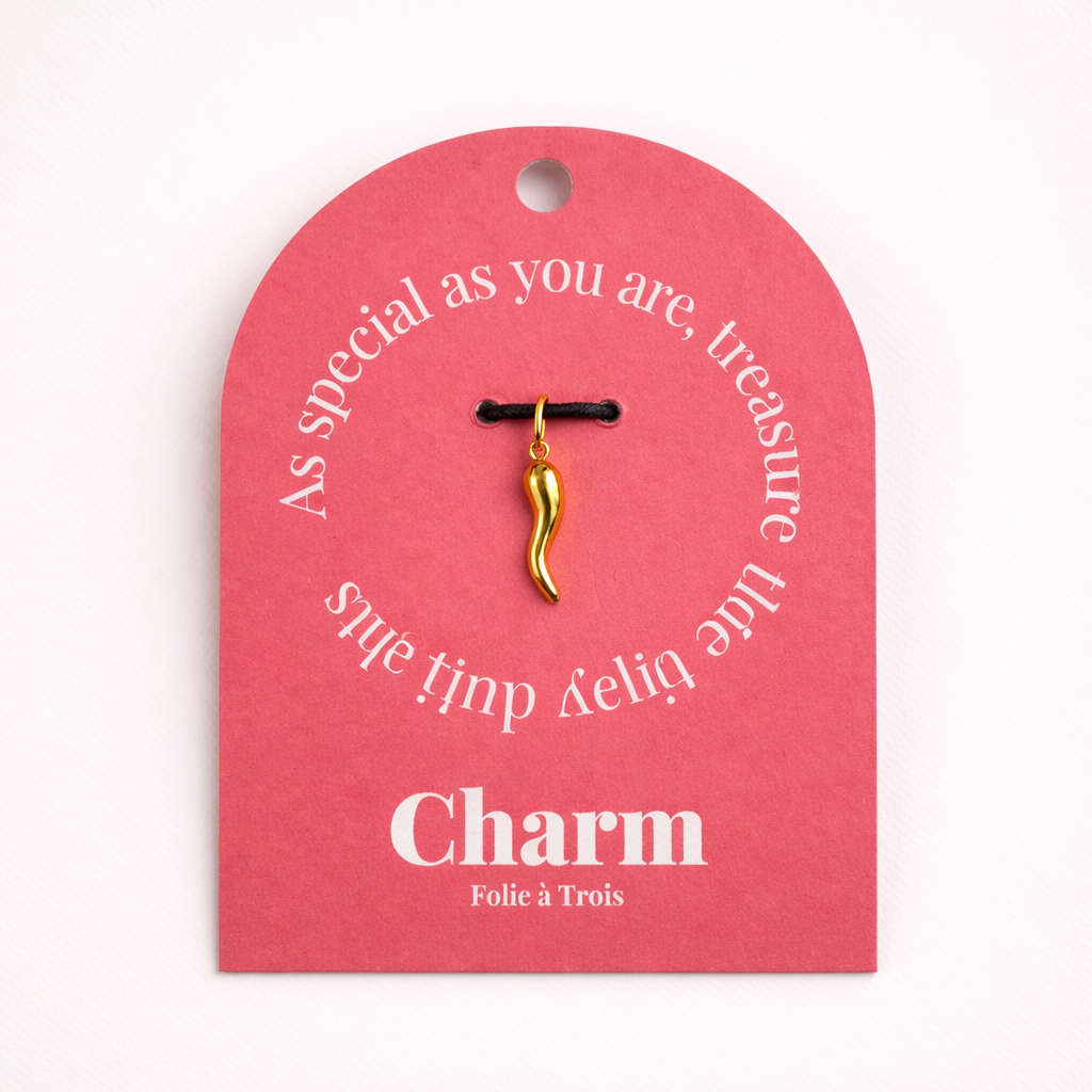 The Gold Chili Pepper CHARM displayed on a coral pink Charm gift tag, the curved gold pendant hanging from a black cord.
