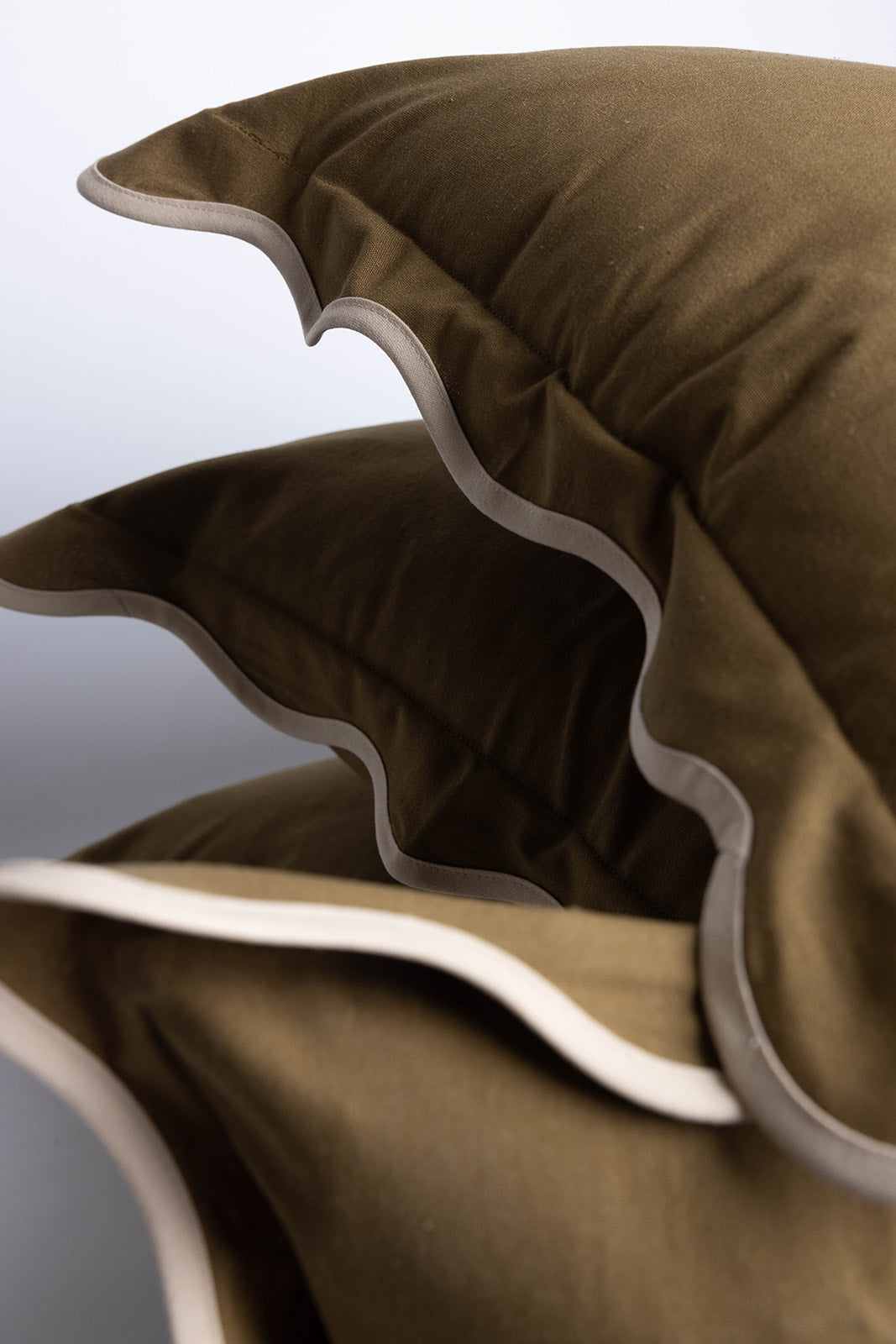Close-up of stacked olive-green Harper Linen cushions showing their soft fabric texture and contrasting scalloped piping edge detail.