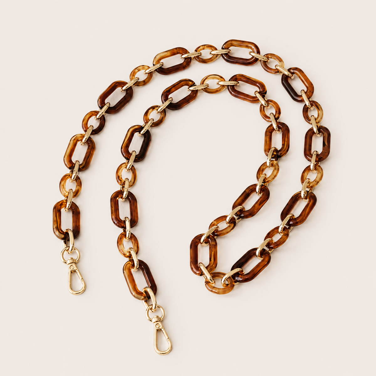 The Hazel LONG PHONE CHAIN in amber tortoiseshell rectangular-link acrylic with gold-tone clasp ends, coiled on a beige background.