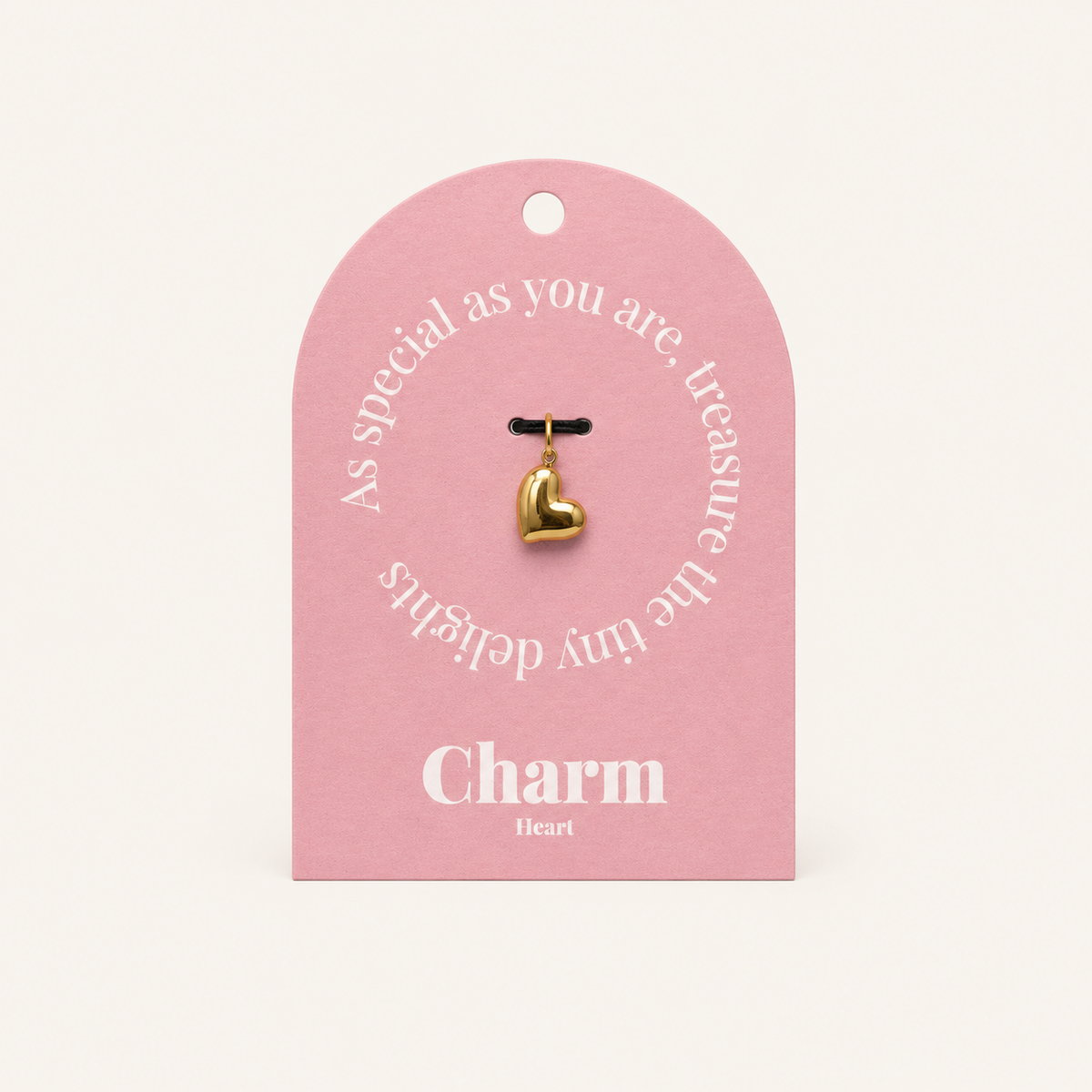 The Heart CHARM displayed on a pink Charm gift tag, the polished gold puffed heart pendant hanging from a black cord.