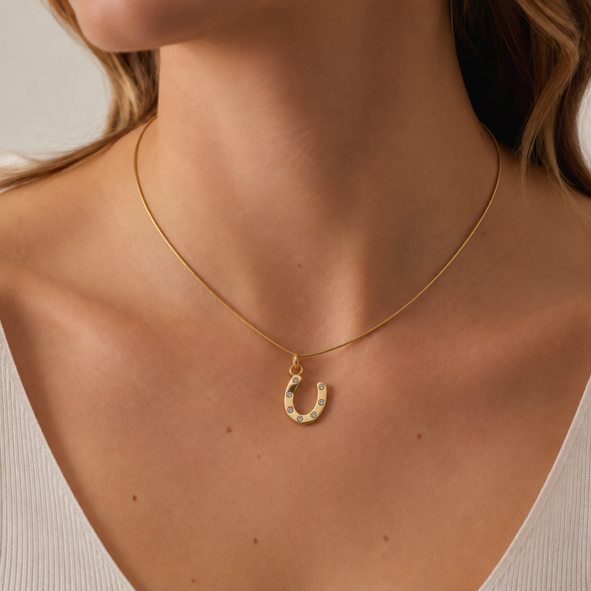 The gold Horseshoe CHARM with clear zircon stones worn on a fine snake chain at the collarbone, styled with a cream ribbed top.