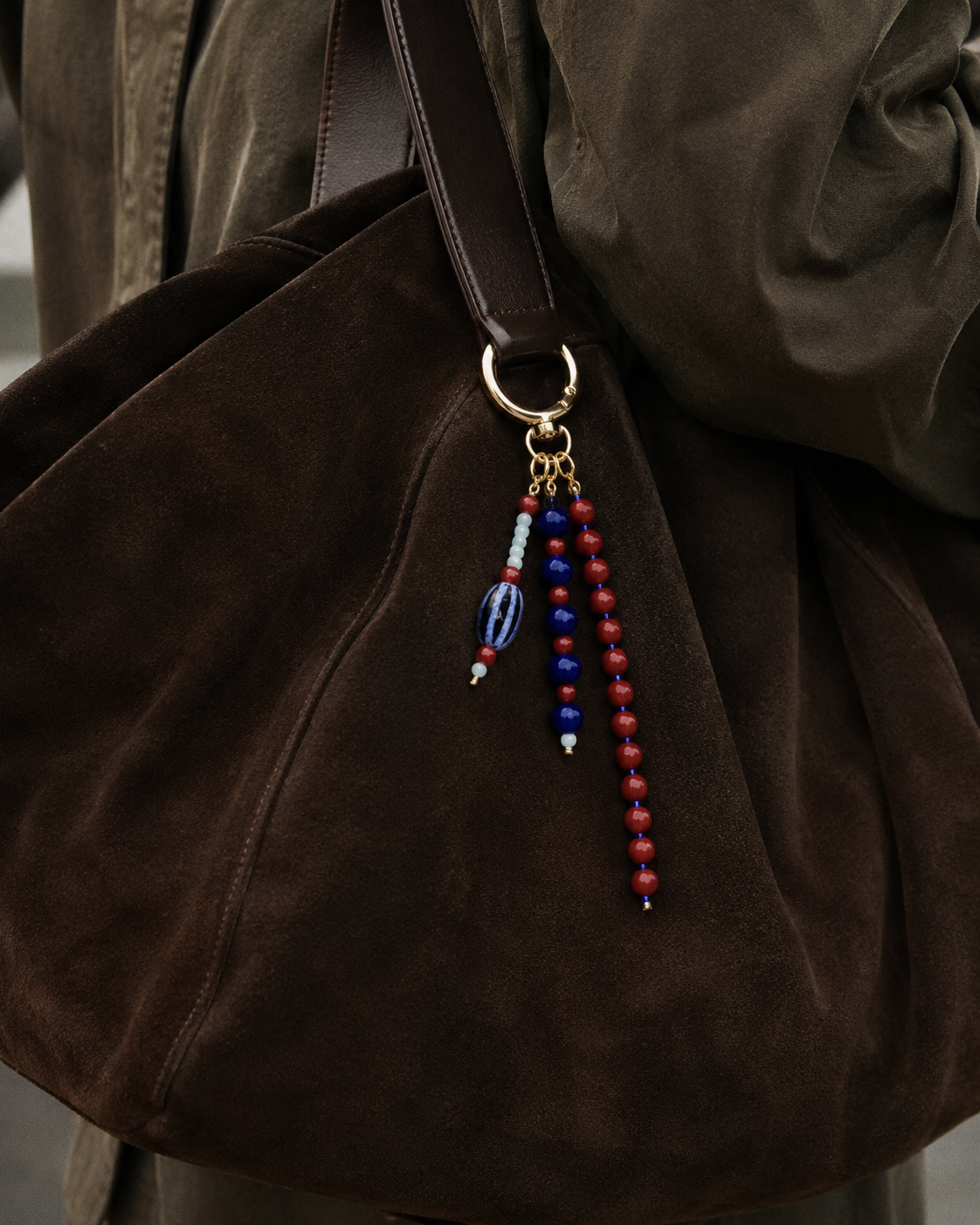 Folie à Trois Isadora BAG CHARM in navy, red and cream beaded strands, clipped to a dark brown suede shoulder bag.