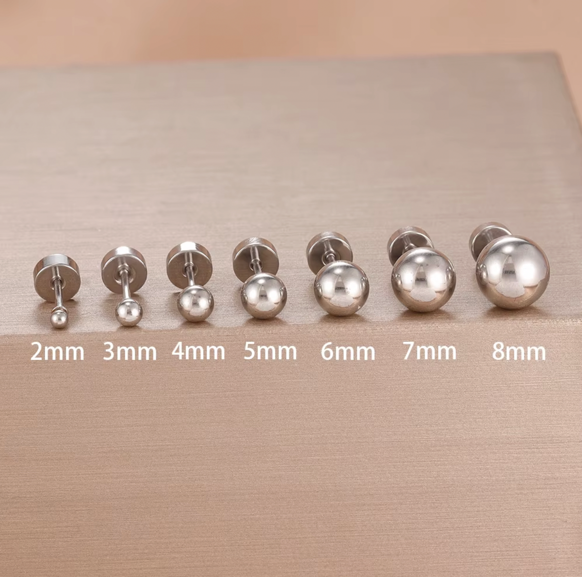 The Jean PIERCING in silver, seven labret studs labeled from 2mm to 8mm displayed on a tan fabric background.