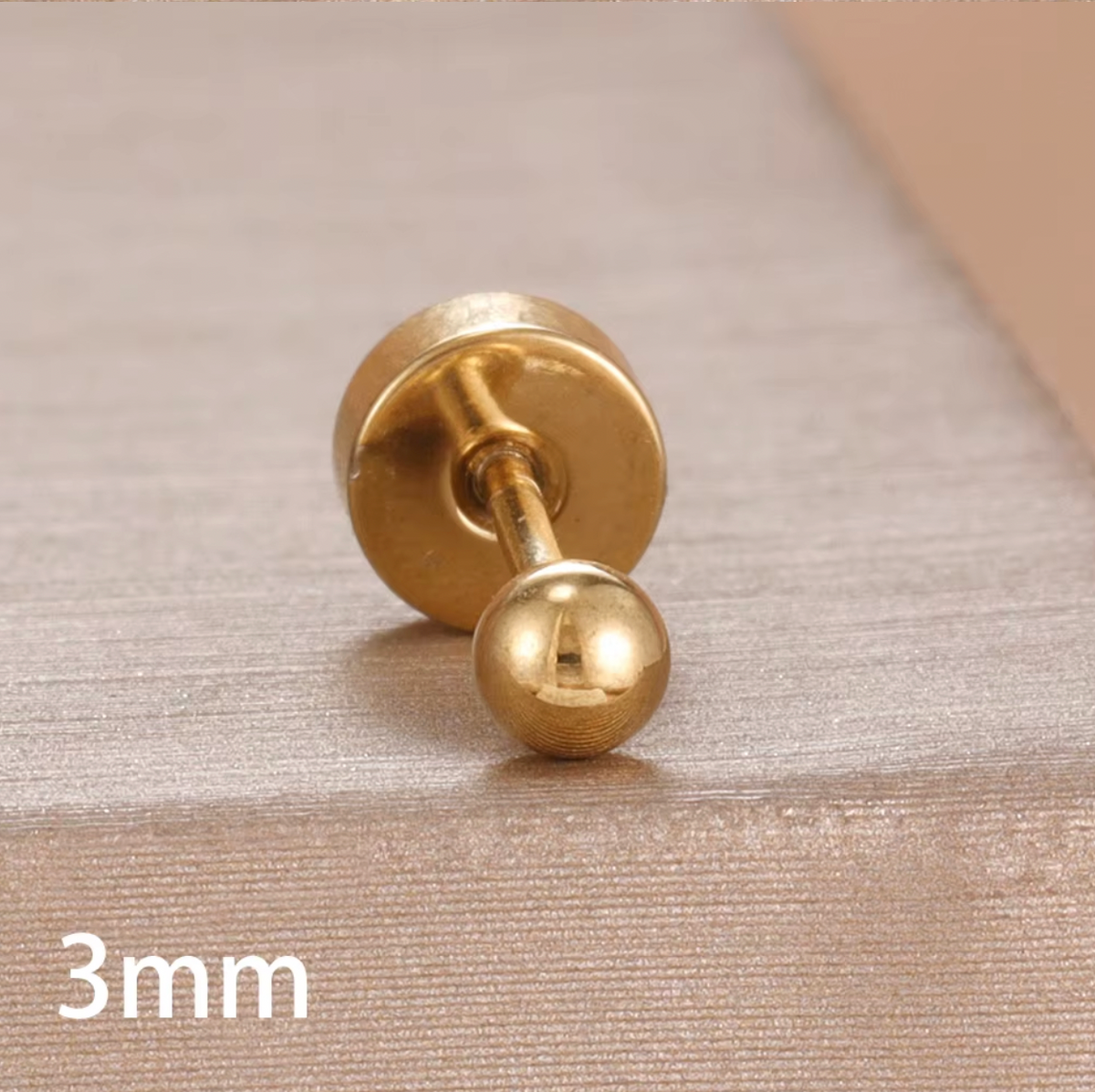 The Jean PIERCING in gold, a single 3mm labret stud shown in close-up on a textured tan surface.