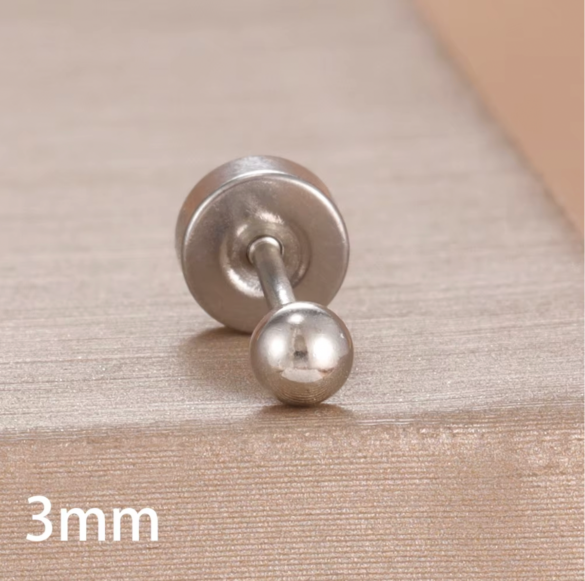 The Jean PIERCING in silver, a single 3mm labret stud shown in close-up on a textured tan surface.