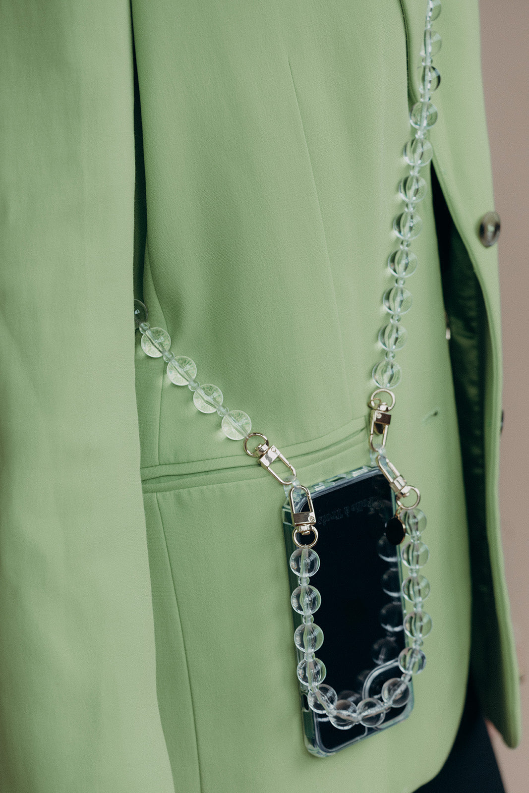 Close-up of a clear glass bead phone chain attached to a phone in a clear case, tucked into the pocket of a sage green blazer.