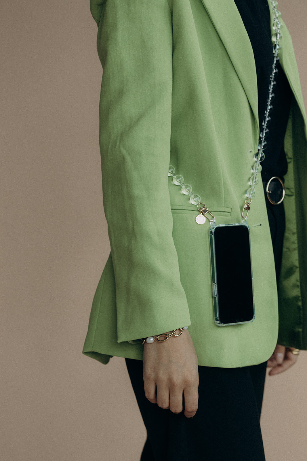 A clear glass bead phone chain draped over the shoulder of a sage green blazer, with a phone tucked in the pocket.