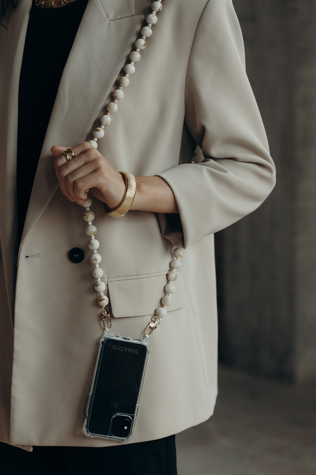 Close-up of a cream and gold marbled bead phone chain held in a hand wearing a ribbed gold bangle, over a beige blazer.