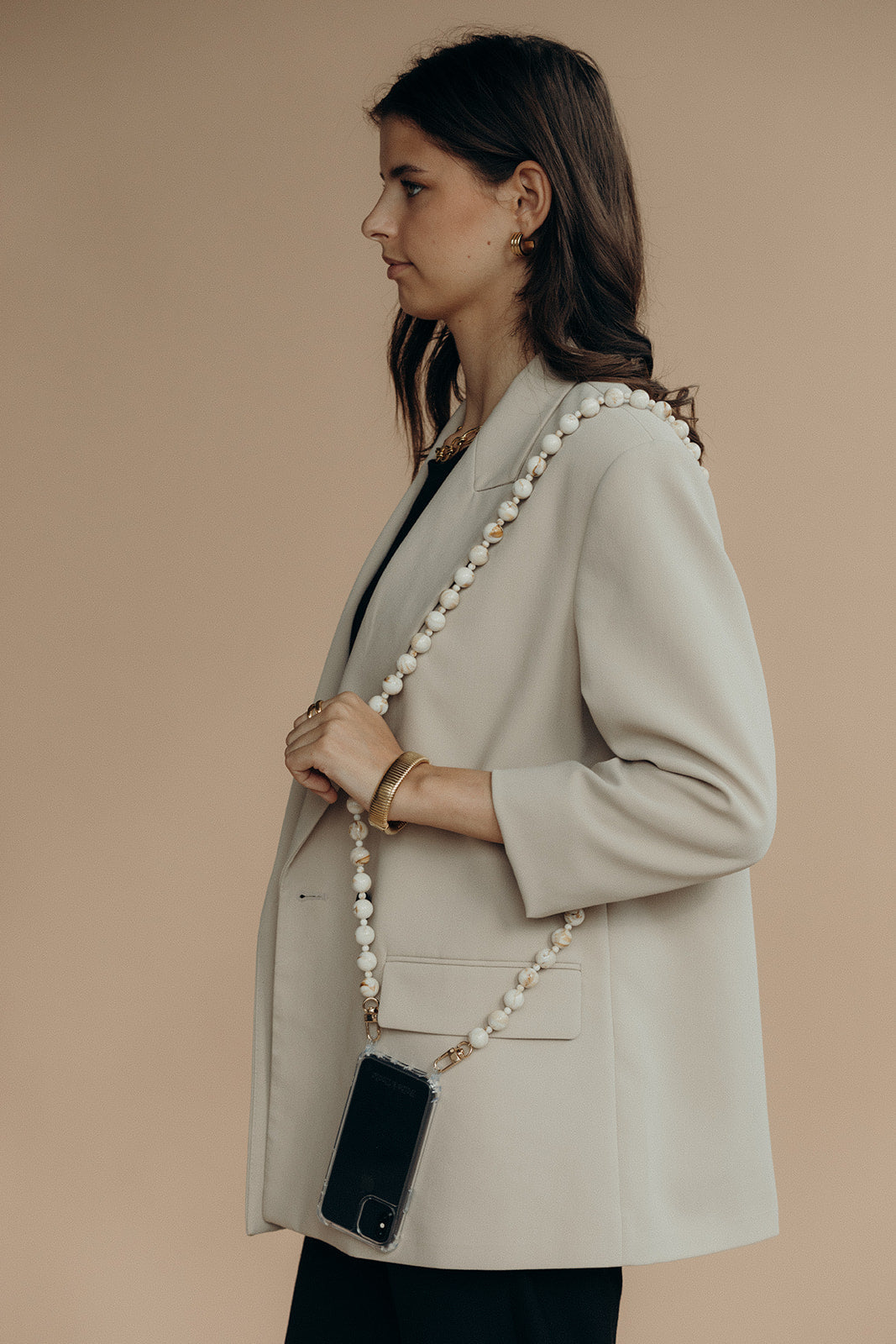 A woman walking while wearing a cream and gold marbled bead phone chain crossbody over a beige blazer.