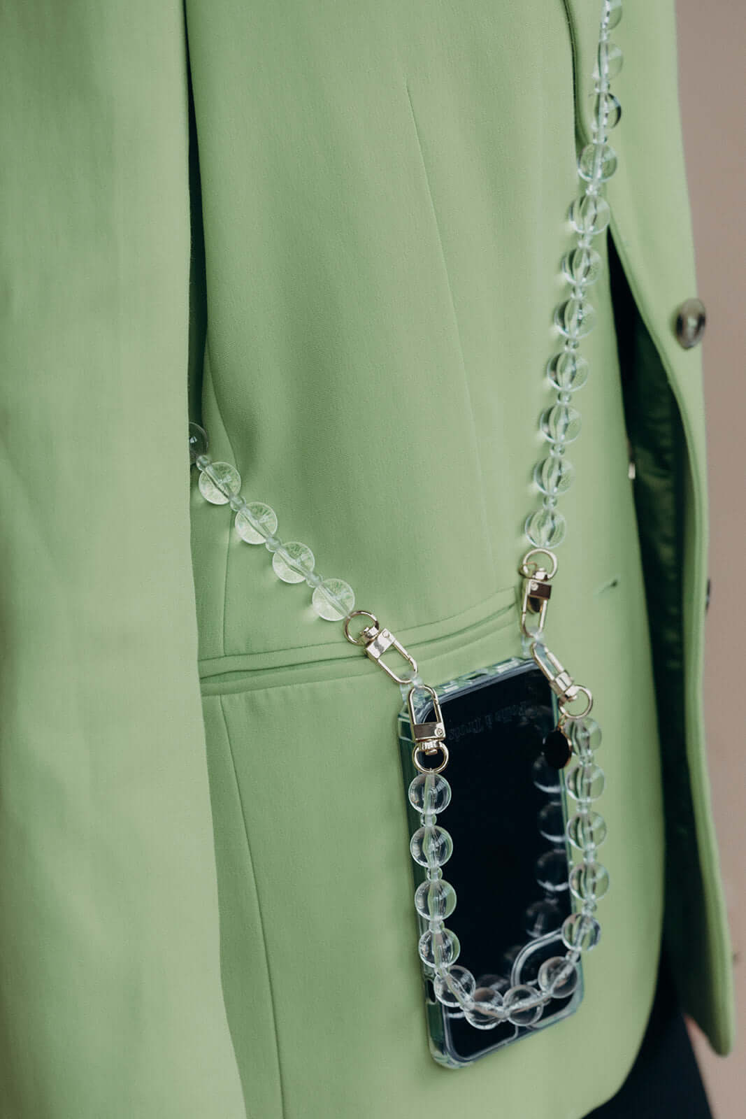 Close-up of a clear glass bead phone strap attached to a phone in a clear case, tucked into the pocket of a sage green blazer.